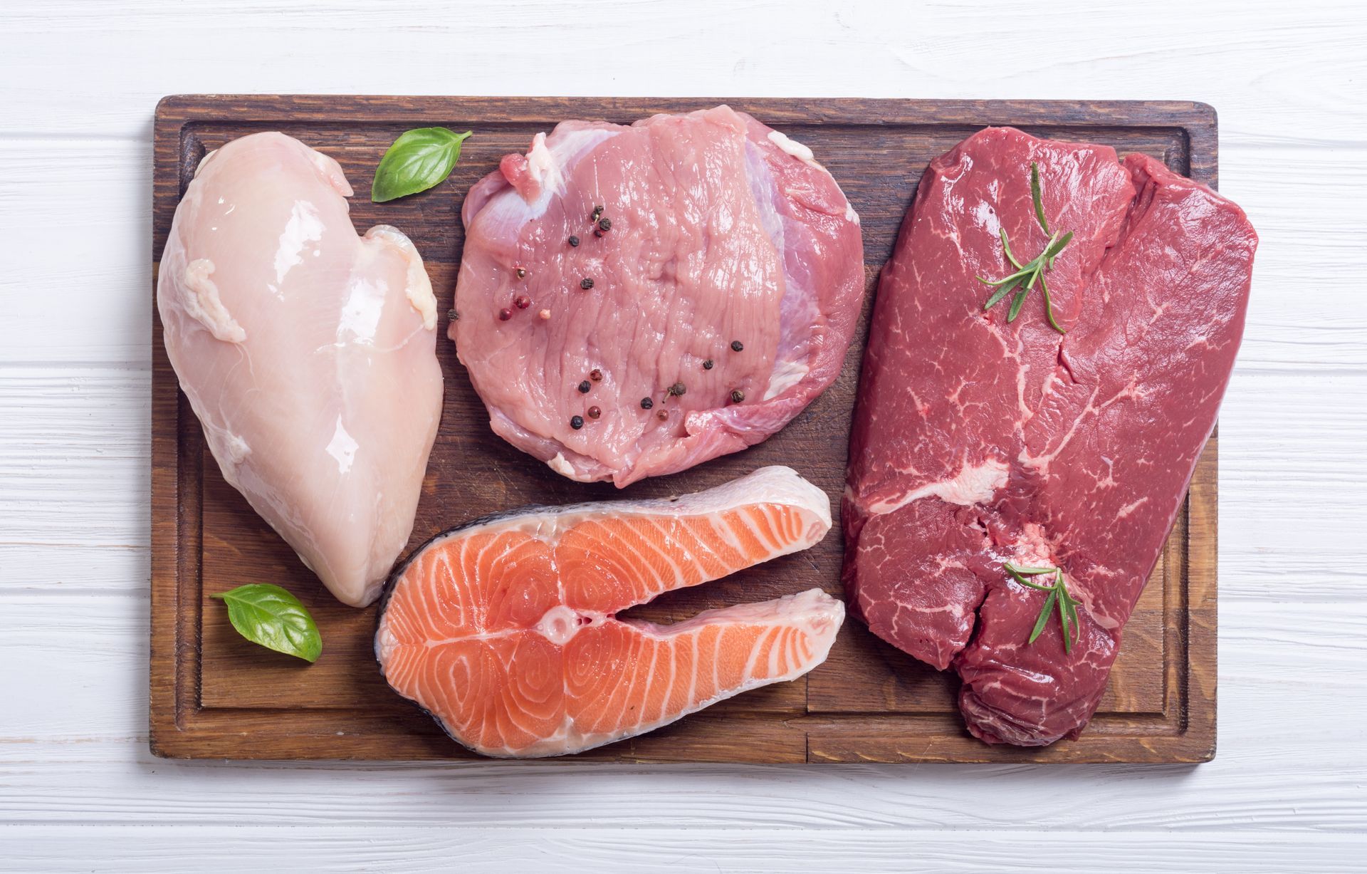 Raw meats and salmon fillet on a wooden cutting board with herb garnish Raw meats and salmon fillet on a wooden cutting board with herb garnish