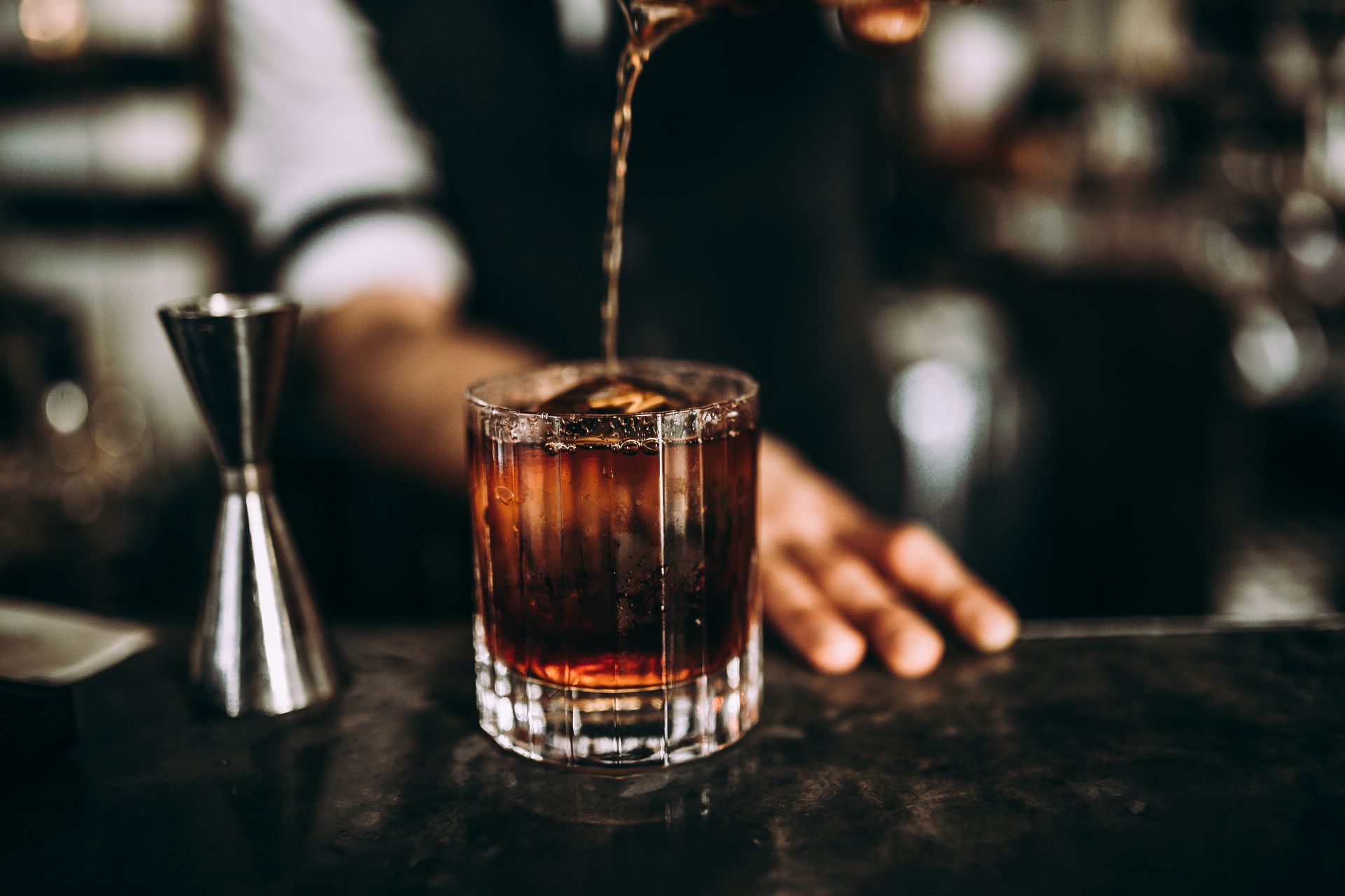 Whiskey being poured into a rocks glass on a bar, with a jigger beside it.