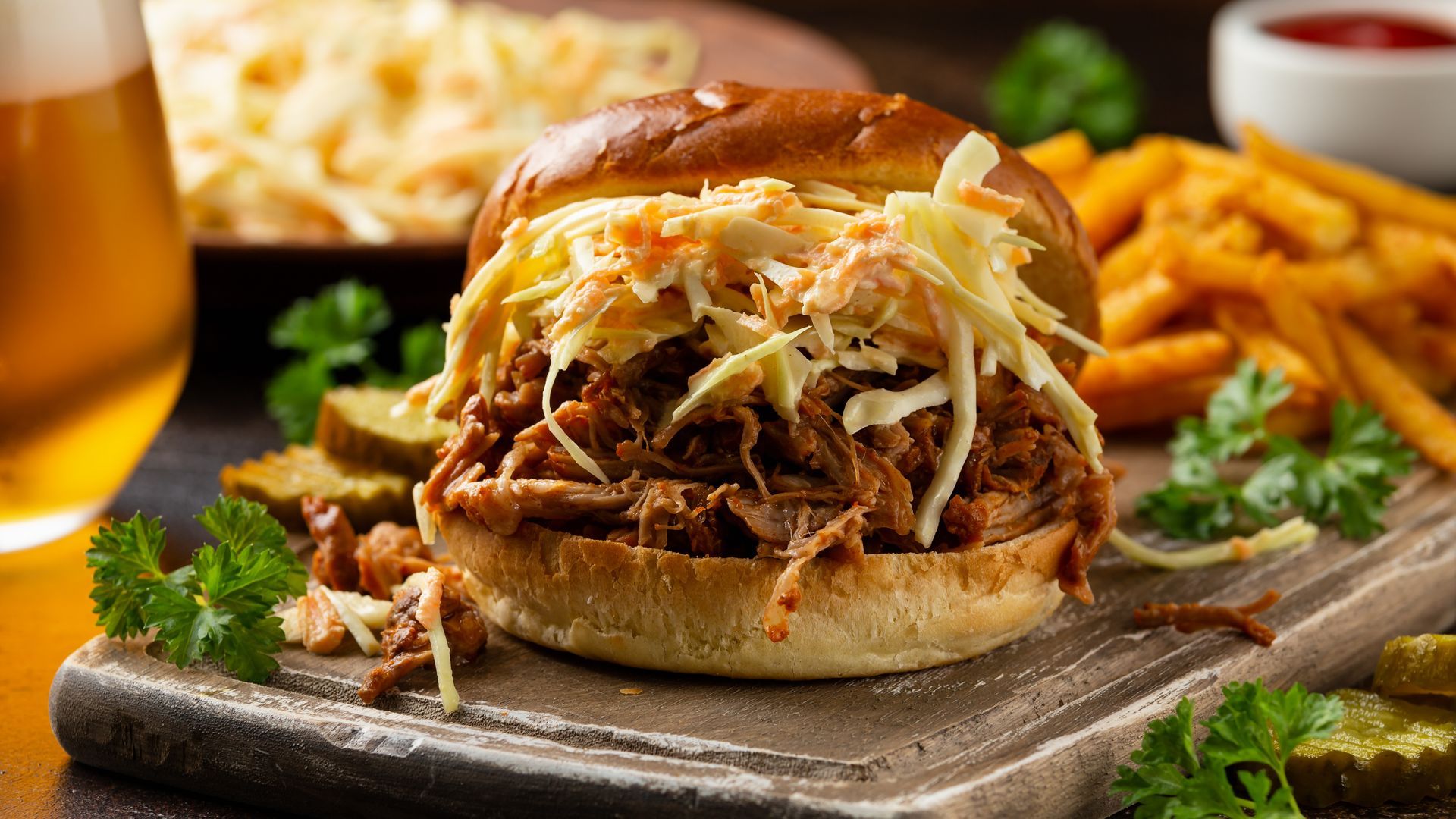 Pulled pork sandwich on a bun with melted cheese, fries, and a beer on a rustic wooden board