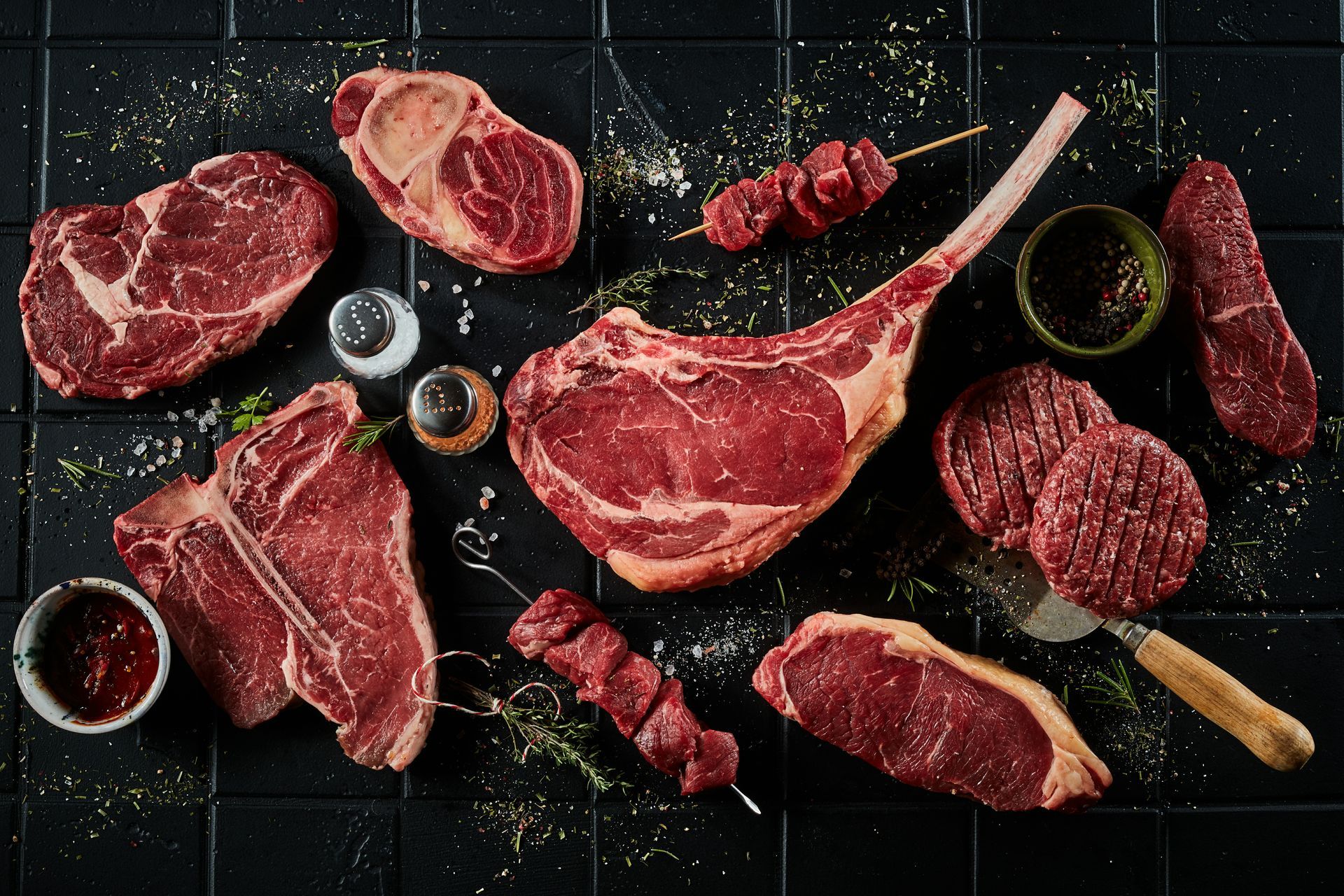 Raw assorted steaks and cuts of beef arranged on a dark slate surface with herbs and seasoning