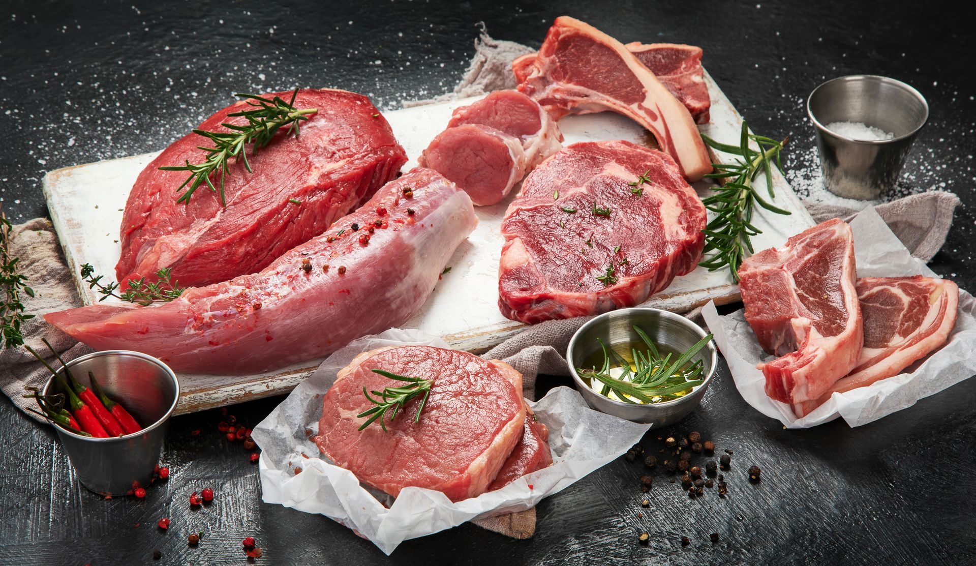 Assorted raw steaks and pork cuts on parchment with herbs and small bowls of seasoning on a dark surface Assorted raw steaks and pork cuts on parchment with herbs and small bowls of seasoning on a dark surface