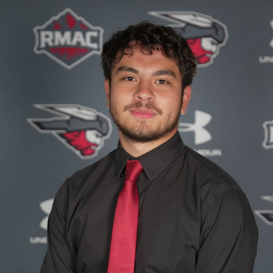 Man wearing a black shirt and red tie, against a backdrop with athletic logos.