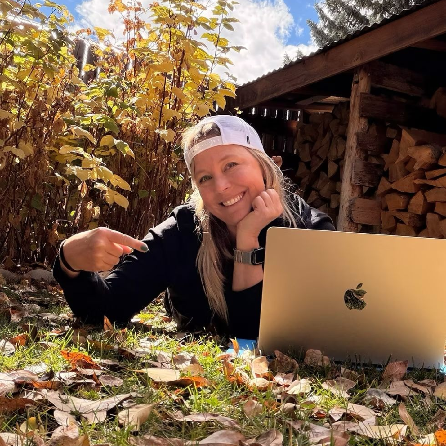 Woman laying on grass with a laptop, pointing. Surrounded by fall leaves and a woodpile.