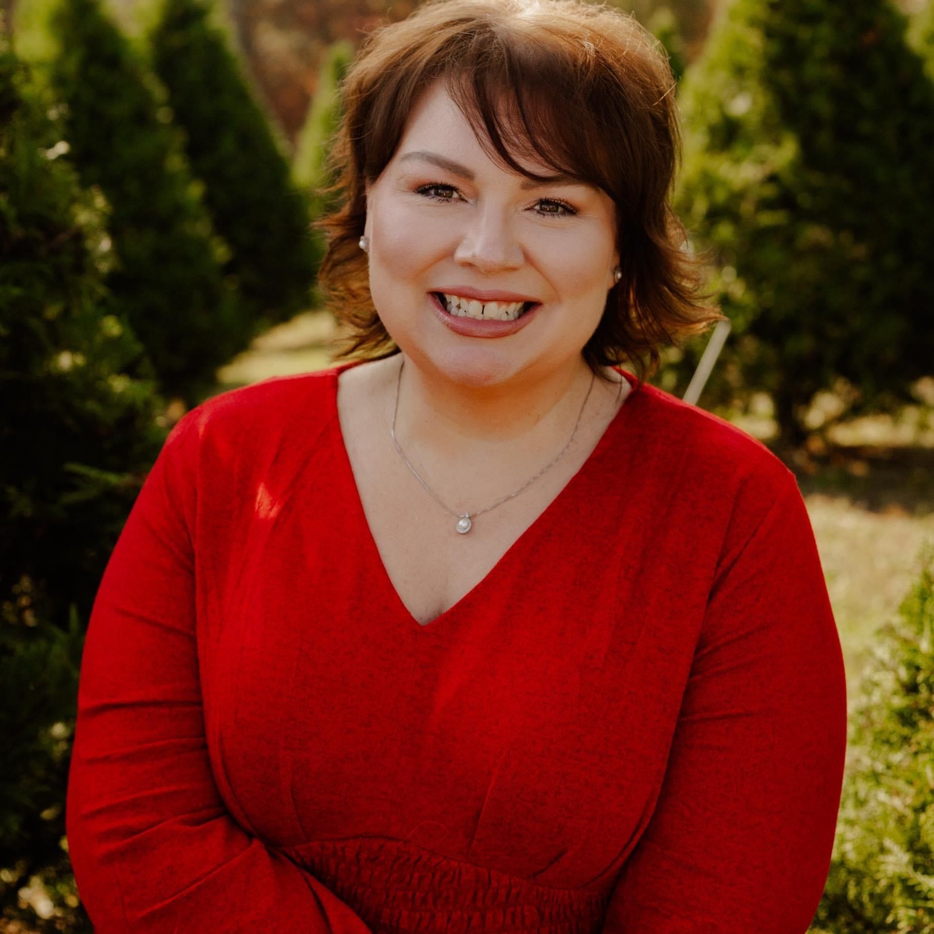 Woman in red shirt smiles outdoors, trees in the background.