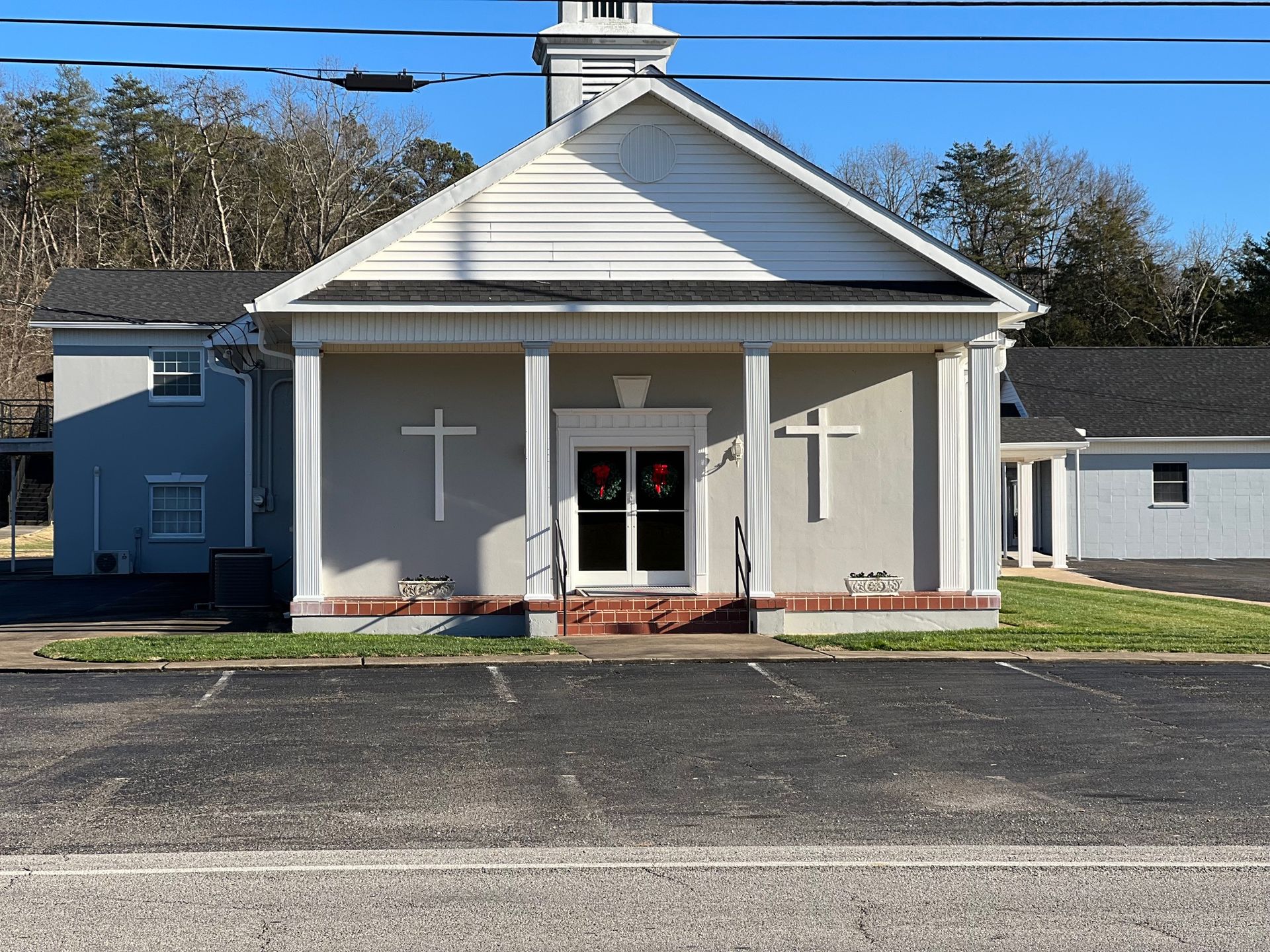 A light gray church with a white steeple, a white cross on the wall, and a black parking lot in front.
