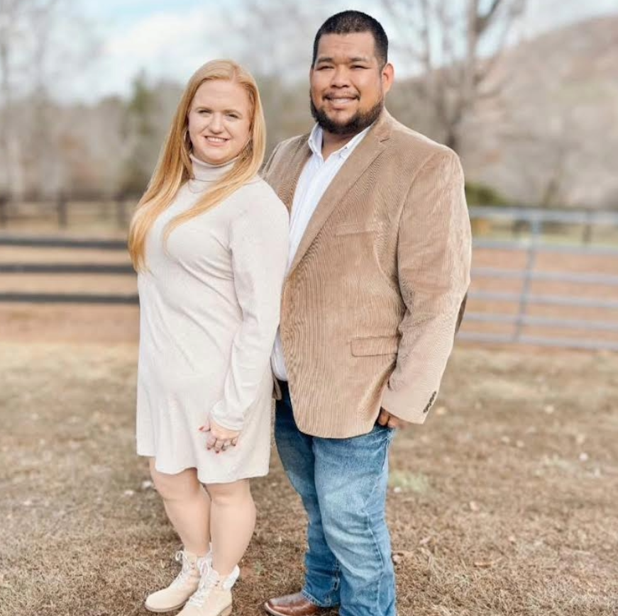 Couple poses outdoors: woman in beige dress, man in corduroy jacket and jeans.