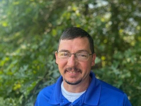Man with glasses smiling, wearing a blue shirt, standing in front of green foliage.