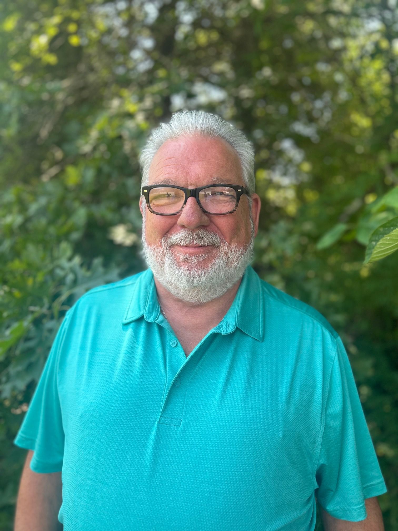 Man with glasses and gray beard smiles, wearing a teal polo shirt, outdoors near green foliage.