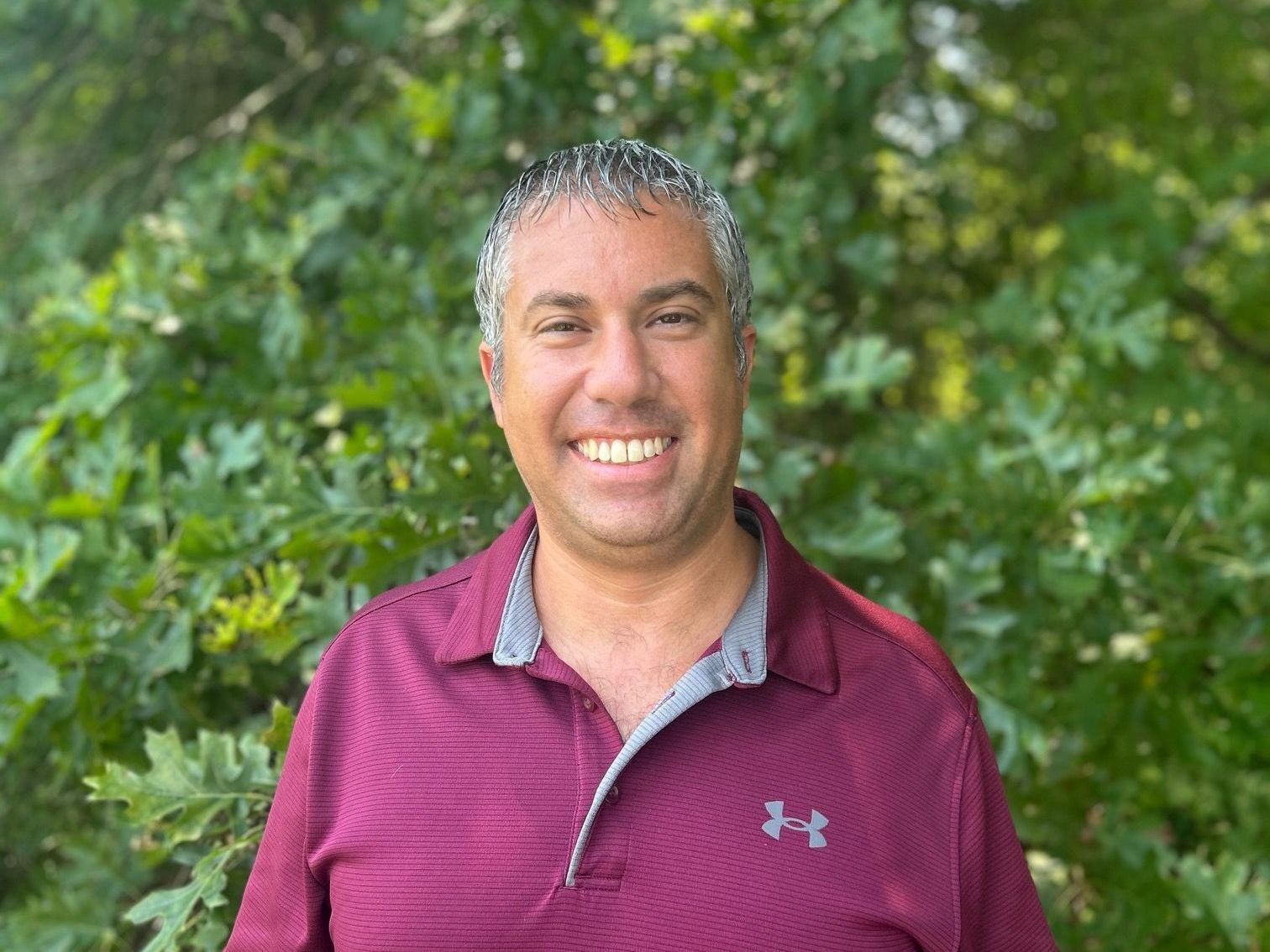 Man with graying hair smiles at the camera while wearing a burgundy Under Armour polo shirt, against a green leafy background.