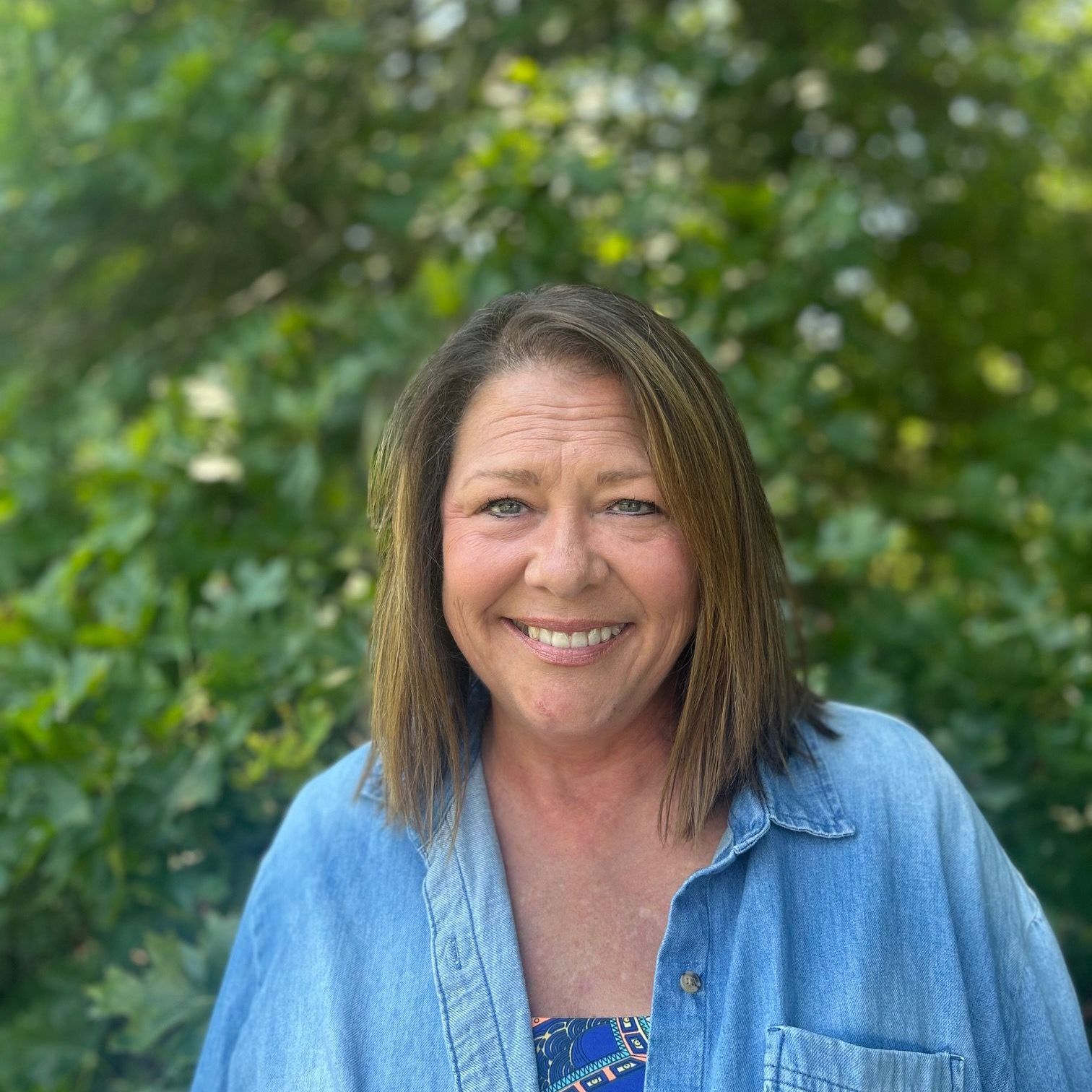 Woman with brown hair smiles in front of green foliage, wearing a blue denim shirt over a patterned top.