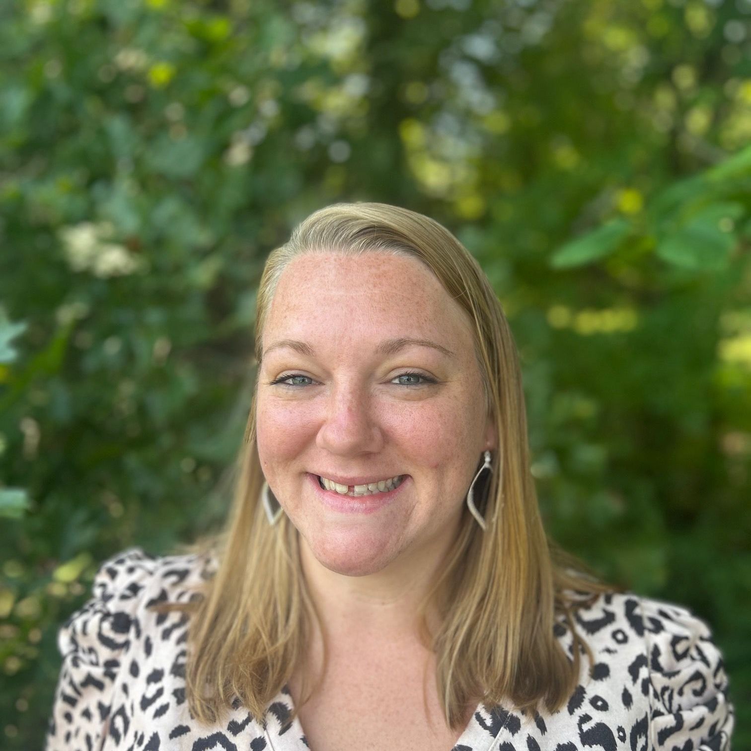 Woman with blonde hair, smiling, wearing animal print top, in front of blurred green foliage.