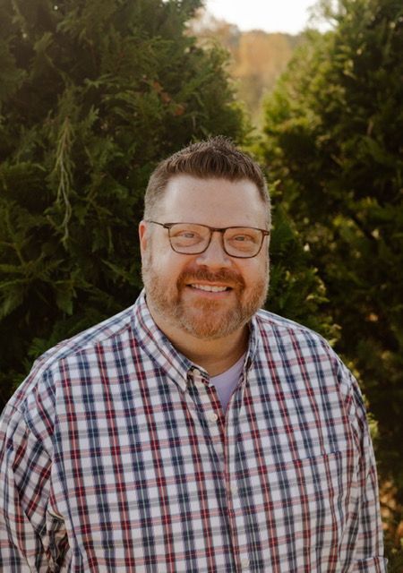 Man with glasses and a beard smiling, wearing a plaid shirt, outdoors with trees in the background.