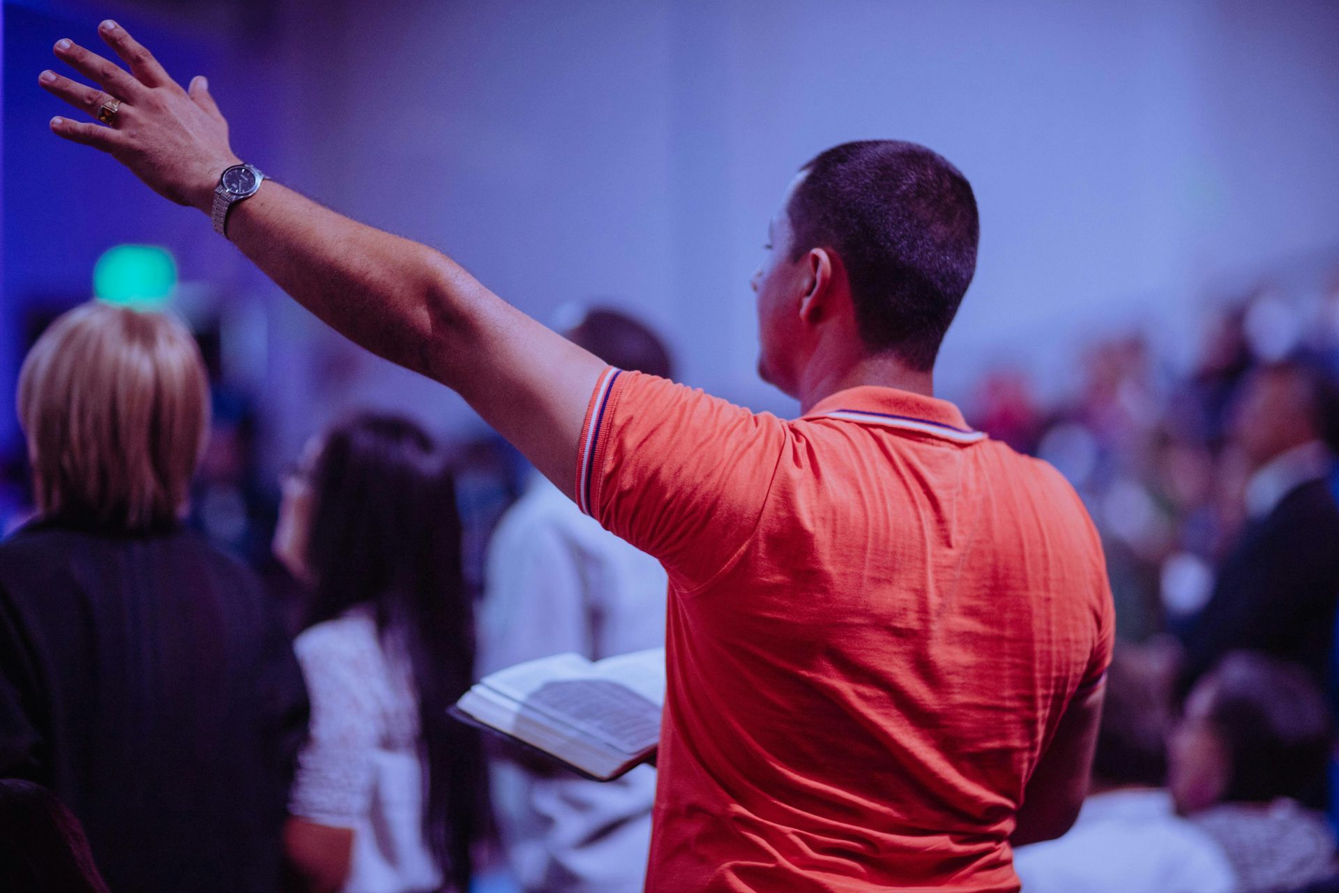 Man with arm raised, holding a book, leading worship in a brightly lit room with people in the background.