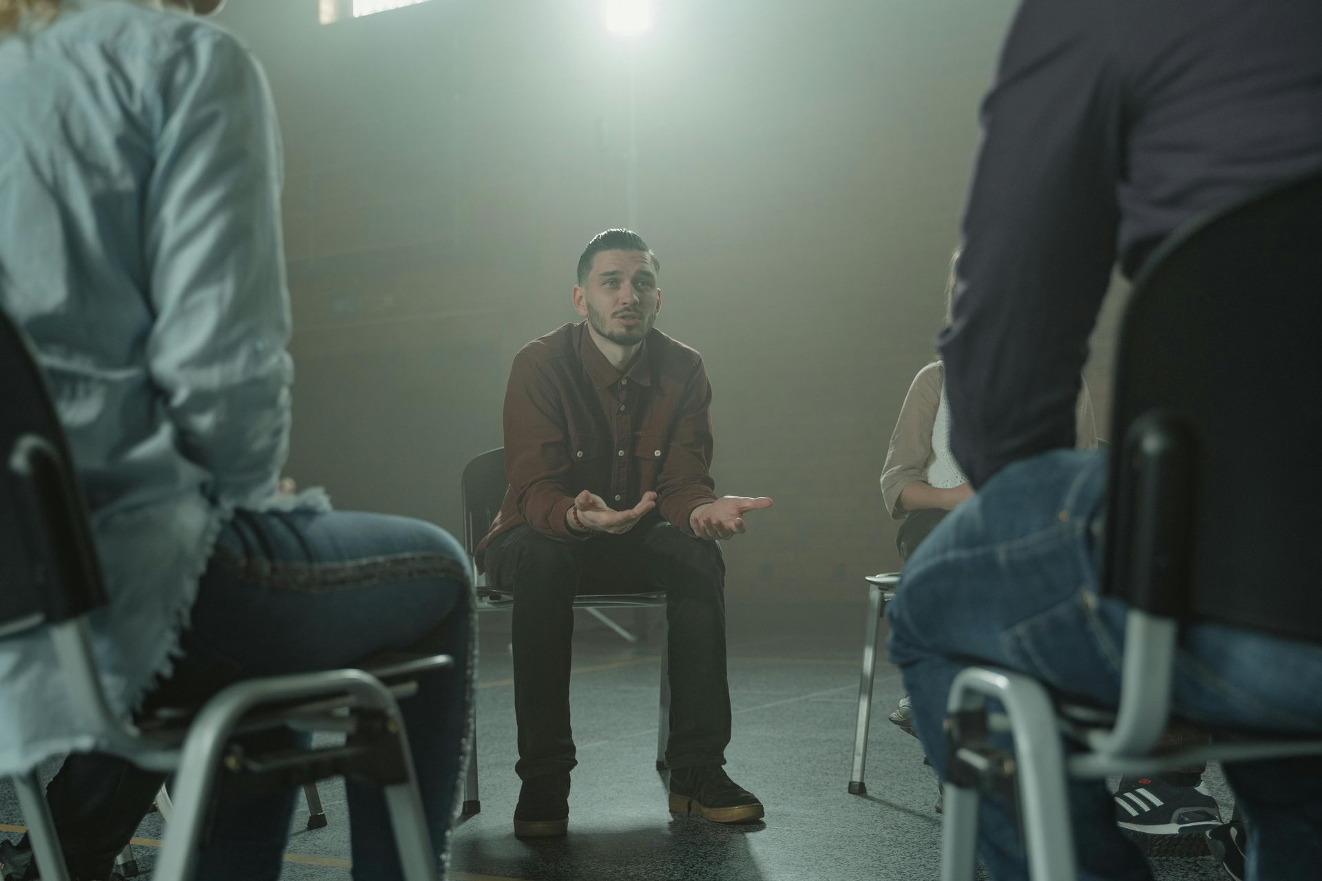 Man in maroon shirt speaking to group in chairs; dimly lit room.