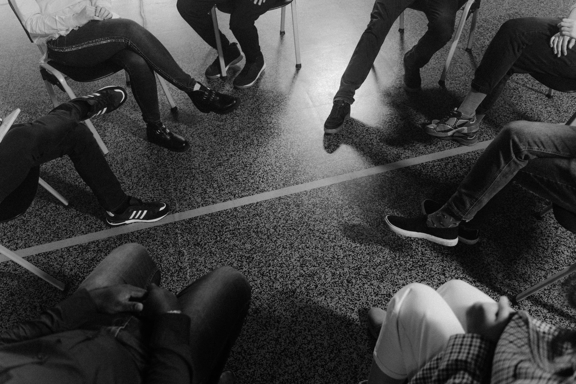 People sit in a circle, legs crossed, near a tape line on a shiny floor. Black and white.