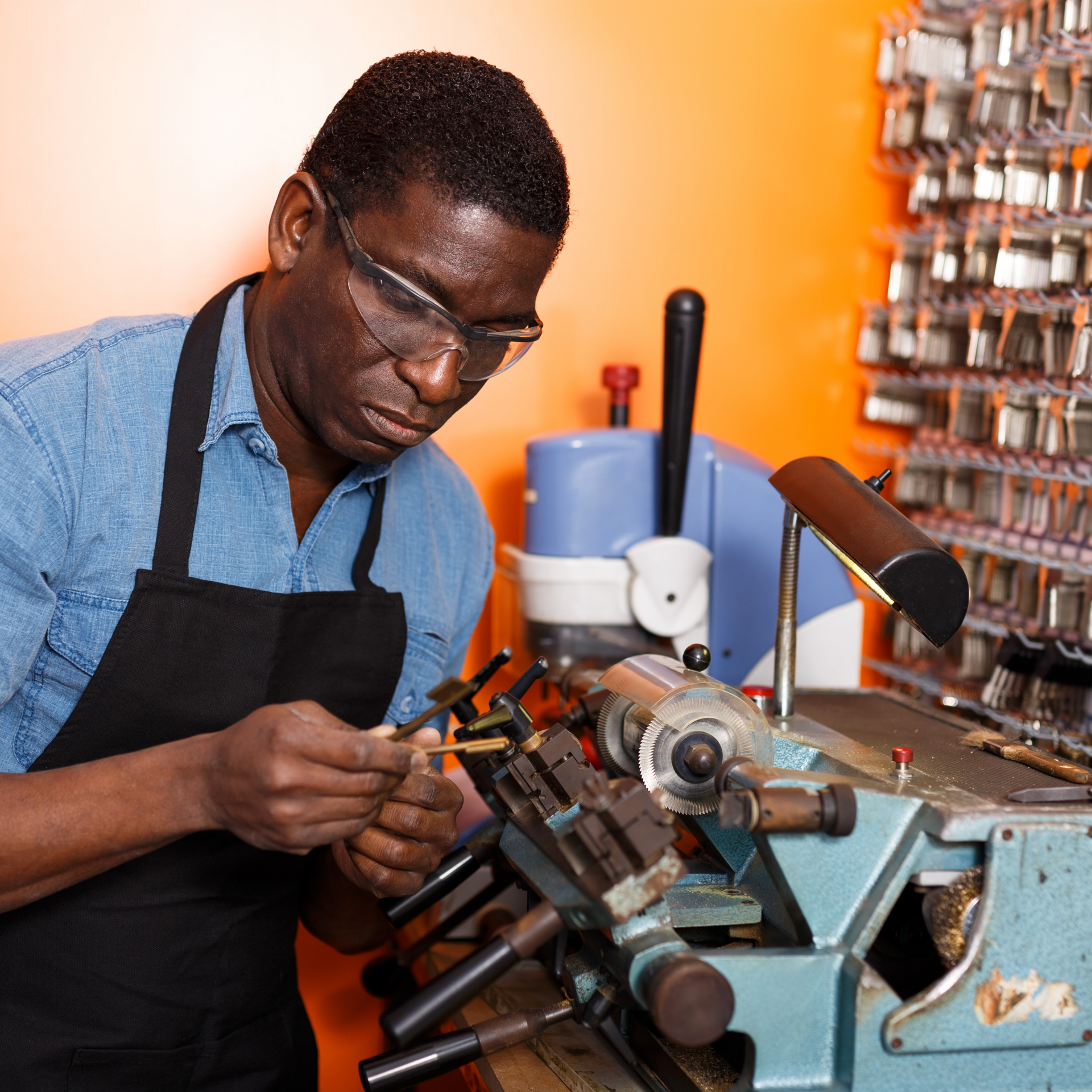 Black man in glasses and apron cutting a key with a key-cutting machine in a workshop.