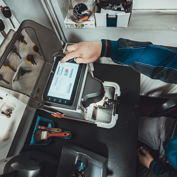 A locksmith using a tablet to program a key-cutting machine in a workshop; a hand points at the screen.