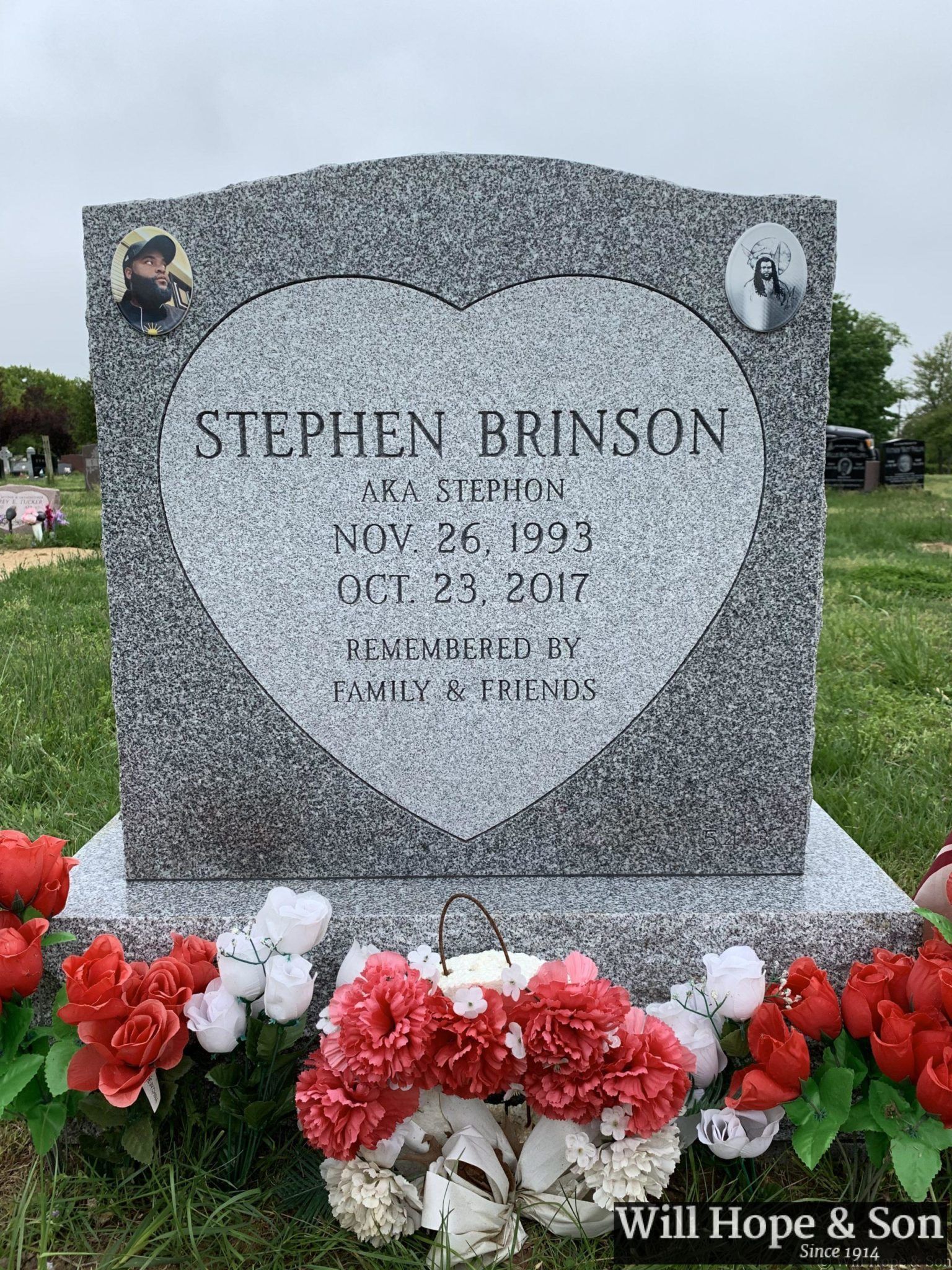 A grey granite headstone for Stephen Brinson with a heart-shaped center, surrounded by artificial flowers in a graveyard.