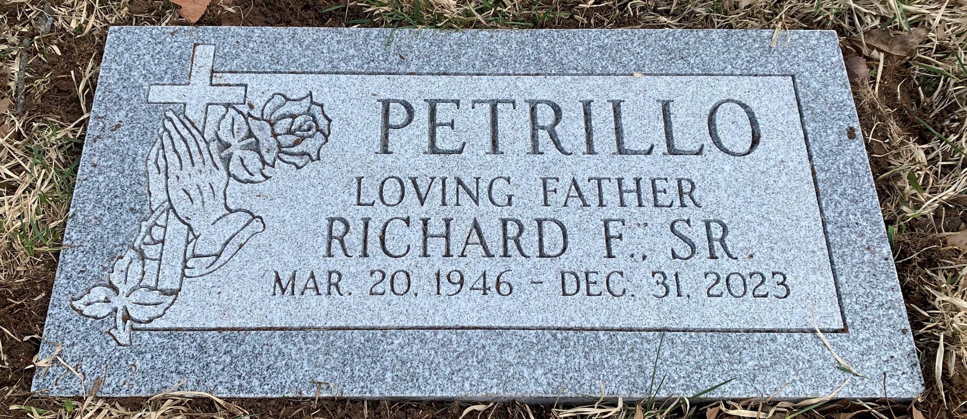 A grey granite headstone for Richard F. Petrillo Sr. (1946–2023) with an engraving of praying hands and a cross.