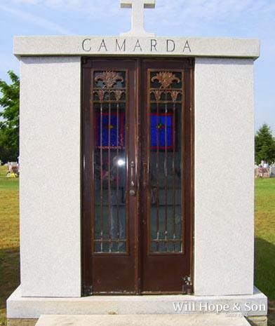 A granite mausoleum for the Camarda family, featuring double doors with metal grilles and a cross on top in a cemetery.