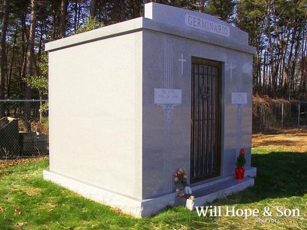 A light-gray granite mausoleum with a dark metal gate, situated on a grassy plot with trees in the background.