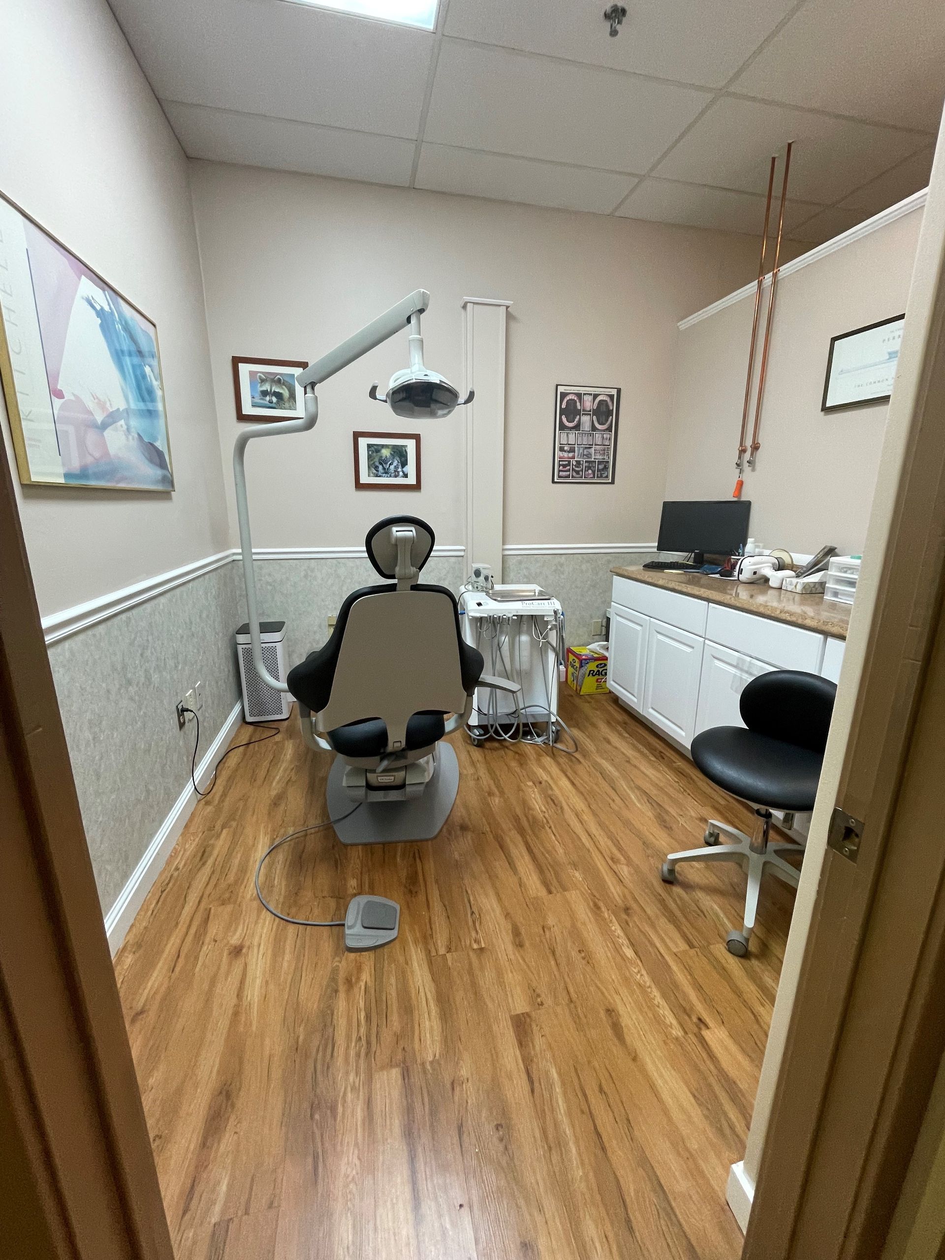 A dental examination room with a chair, light, and cabinets. Wood flooring and beige walls.