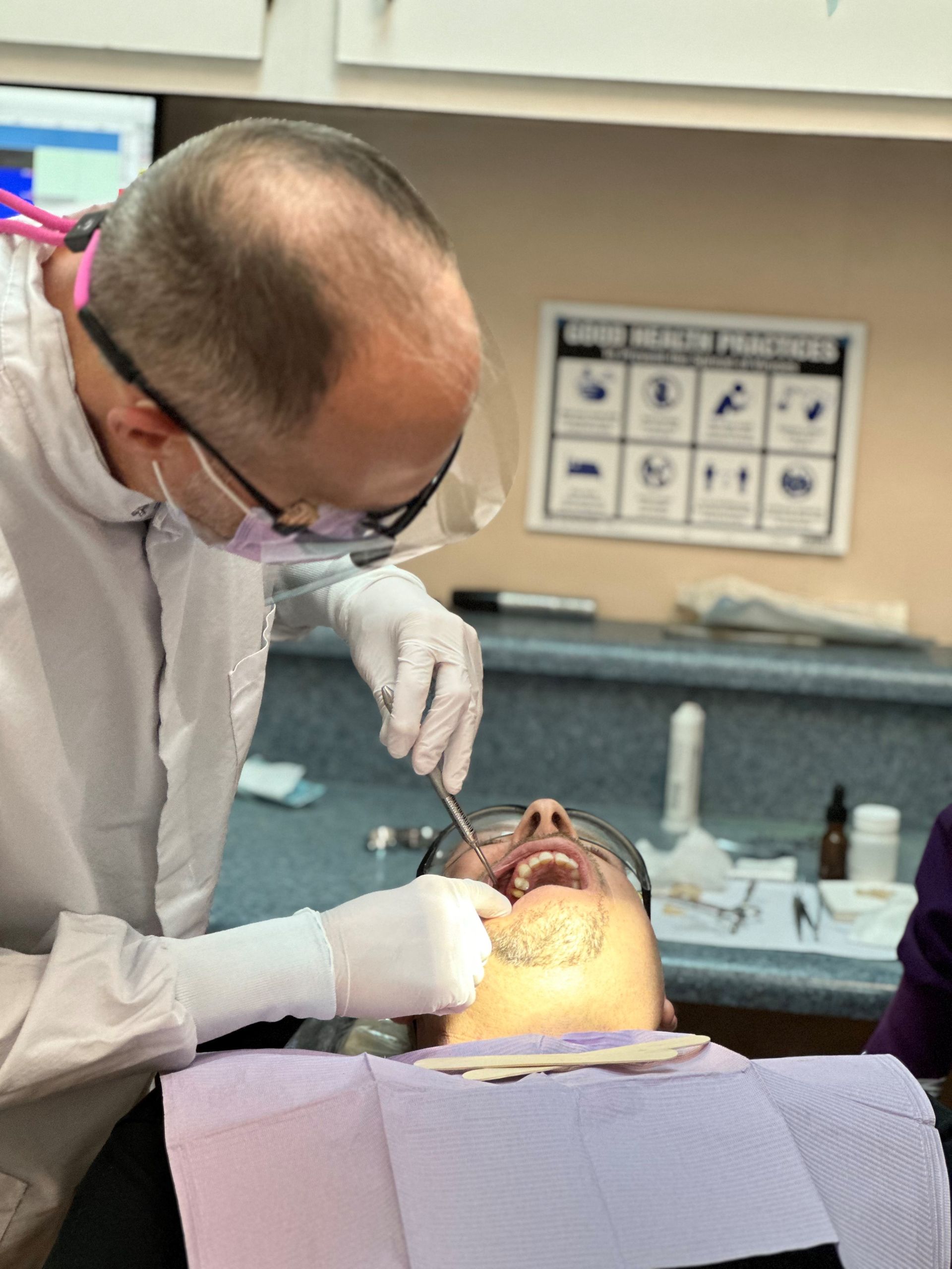 Dentist examining patient's teeth in a dental chair. Dentist wearing gloves, glasses, and a mask, working with tools.