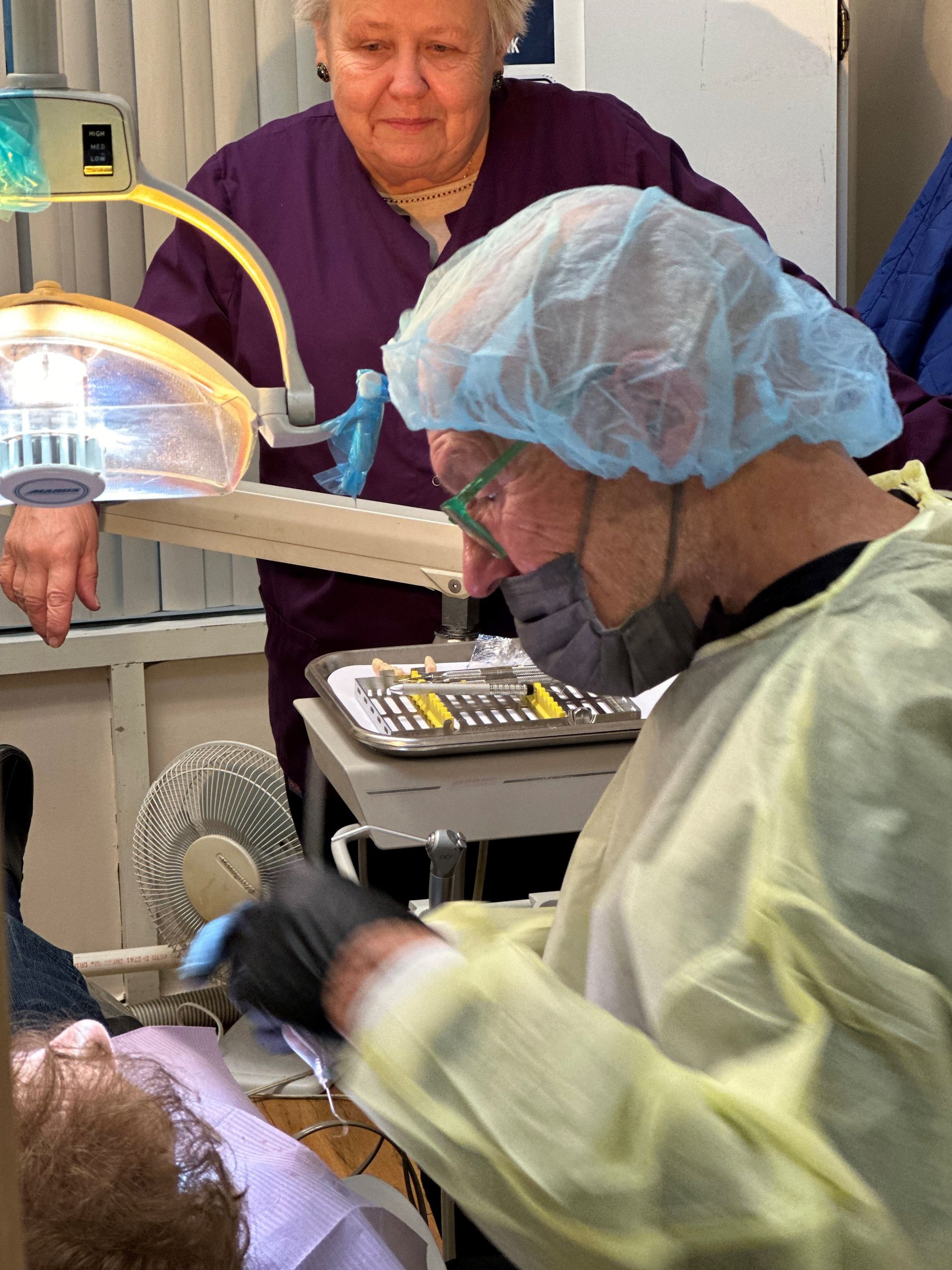 Dentist in protective gear examines patient's mouth, assistant watches. Bright dental lamp.