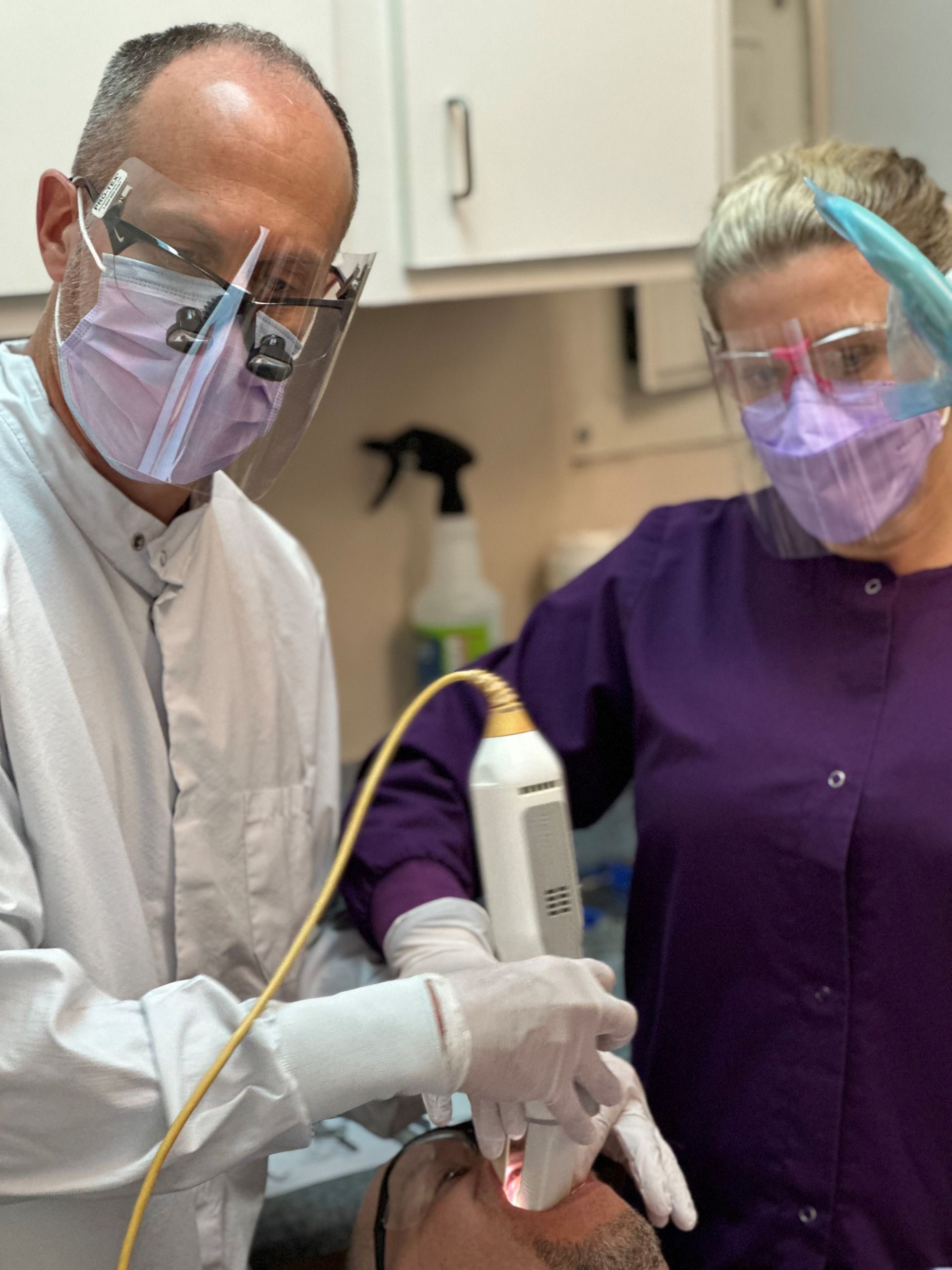 Dentist and assistant using a scanner on a patient in a dental office. Both wear masks, face shields, and gloves.