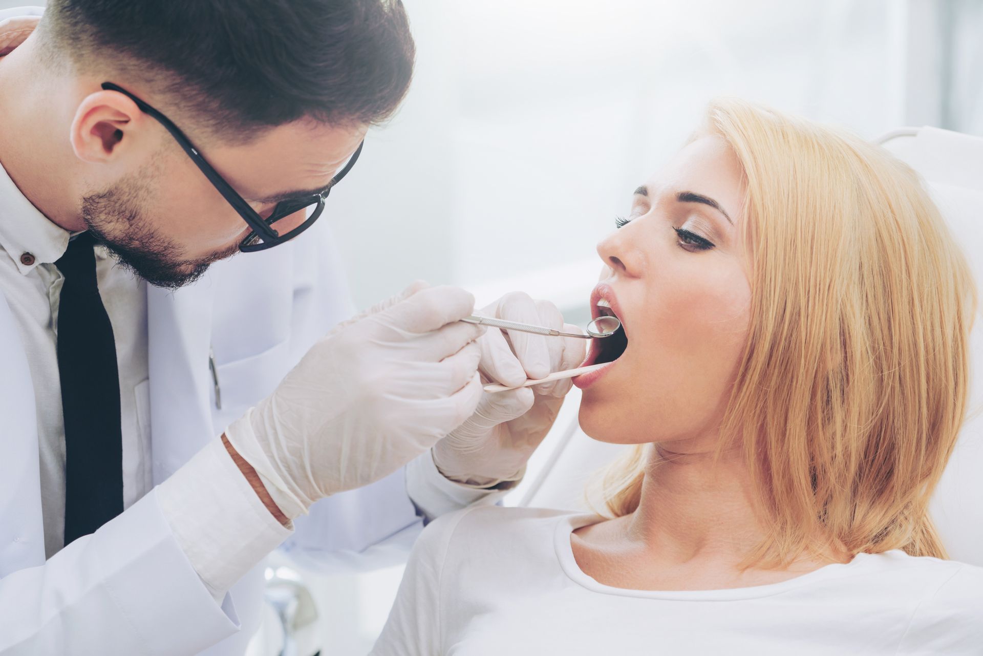 Dentist using dental mirror to examine female patient's teeth during oral checkup.