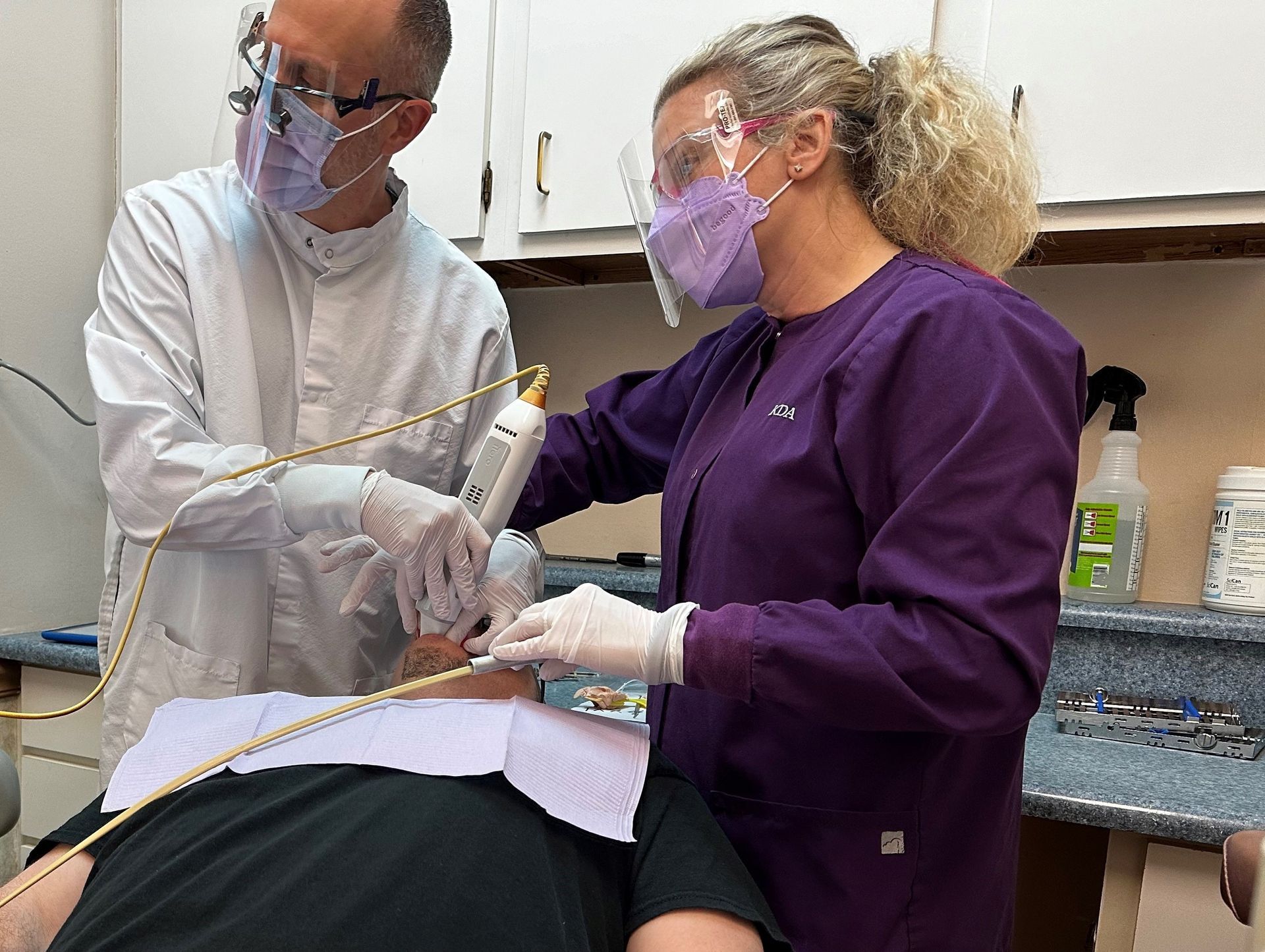 Dentist and assistant in masks/face shields treating a patient in a dental chair.