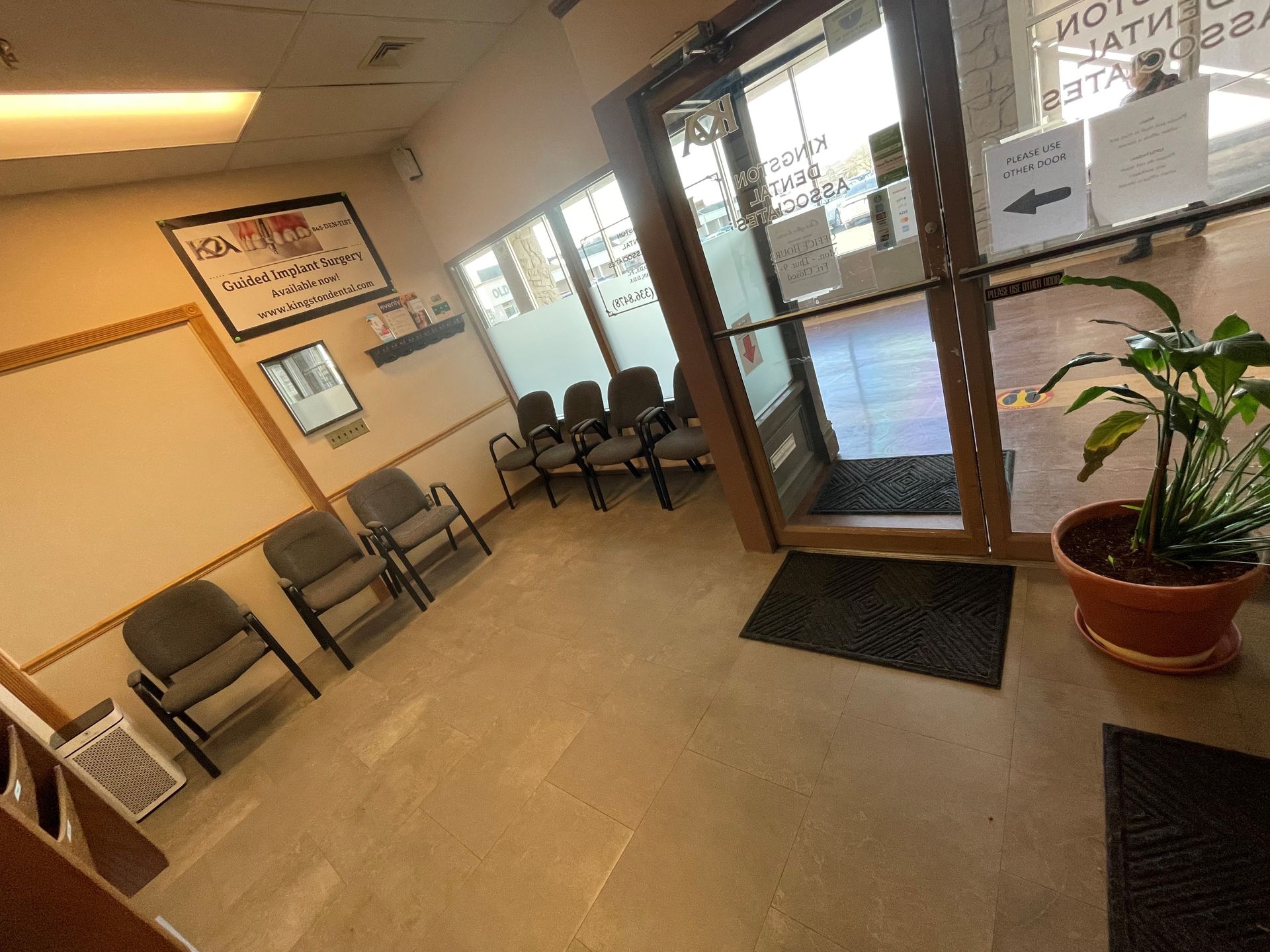 Interior view of a waiting room with chairs, potted plant, and a glass door leading outside.