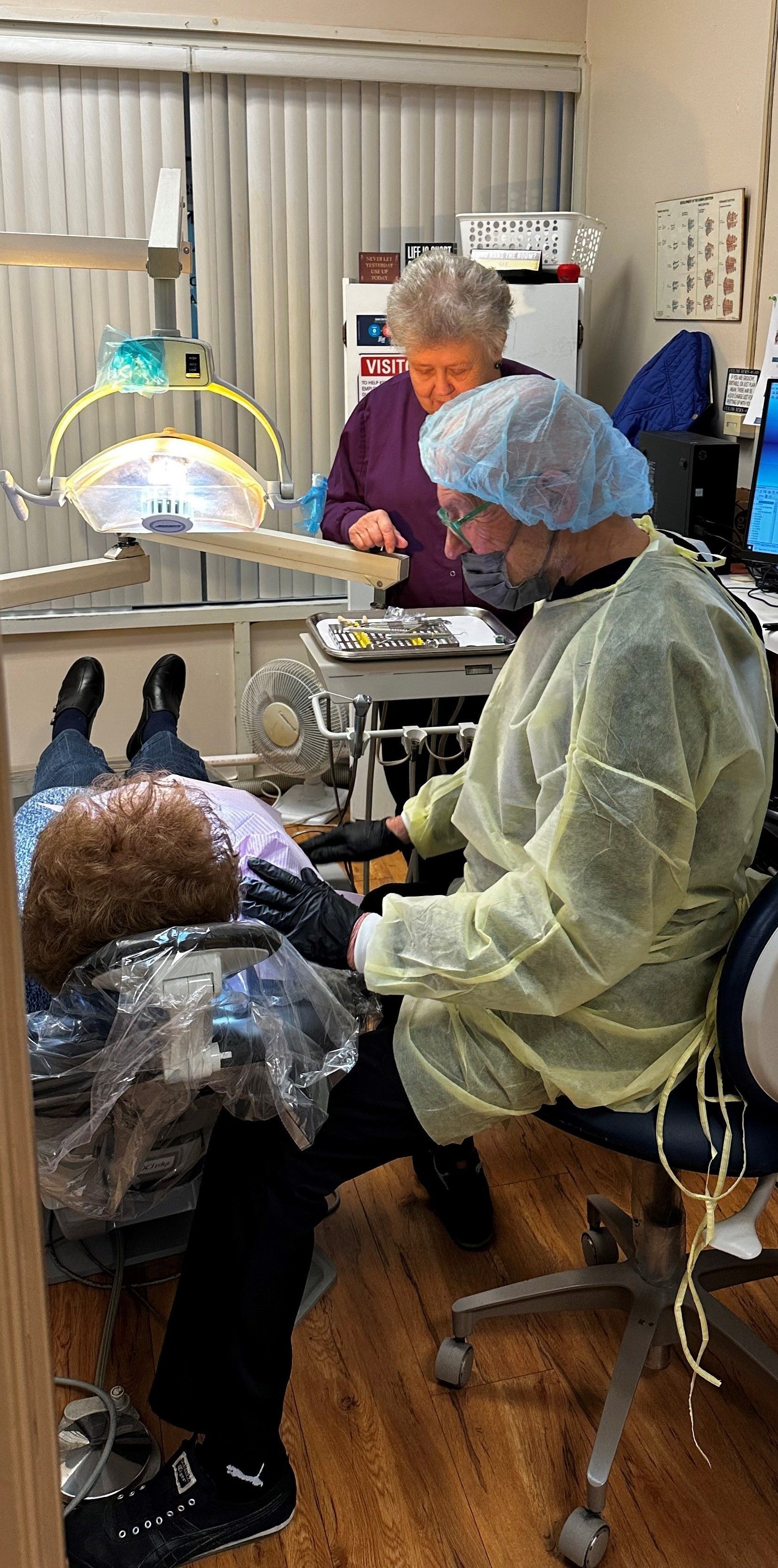 Dentist in protective gear examining a patient in a dental office, with assistant present.