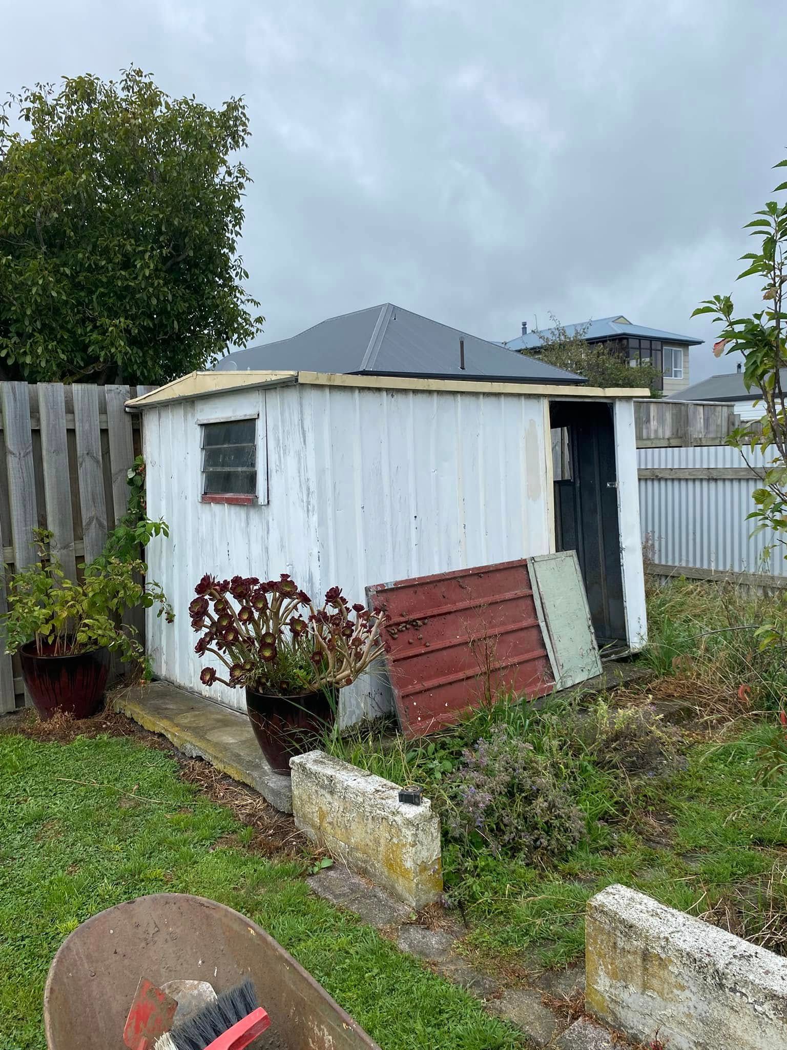 White shed in a backyard with a cloudy sky, next to a fence and overgrown plants.