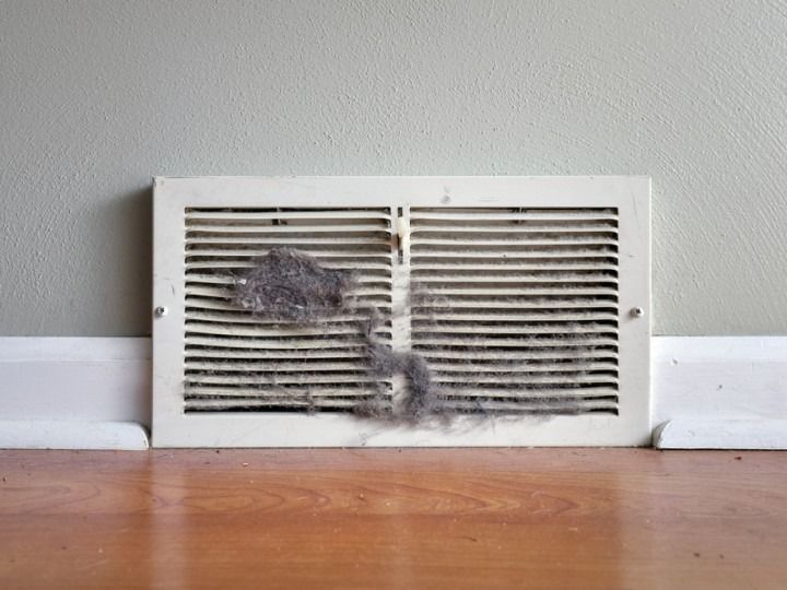 Dirty air vent with visible dust and debris on a hardwood floor against a light green wall.