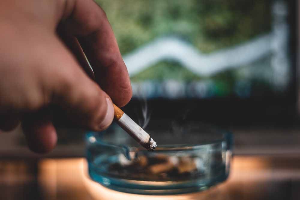 Hand holding a lit cigarette over a glass ashtray with cigarette butts. Blurred background.