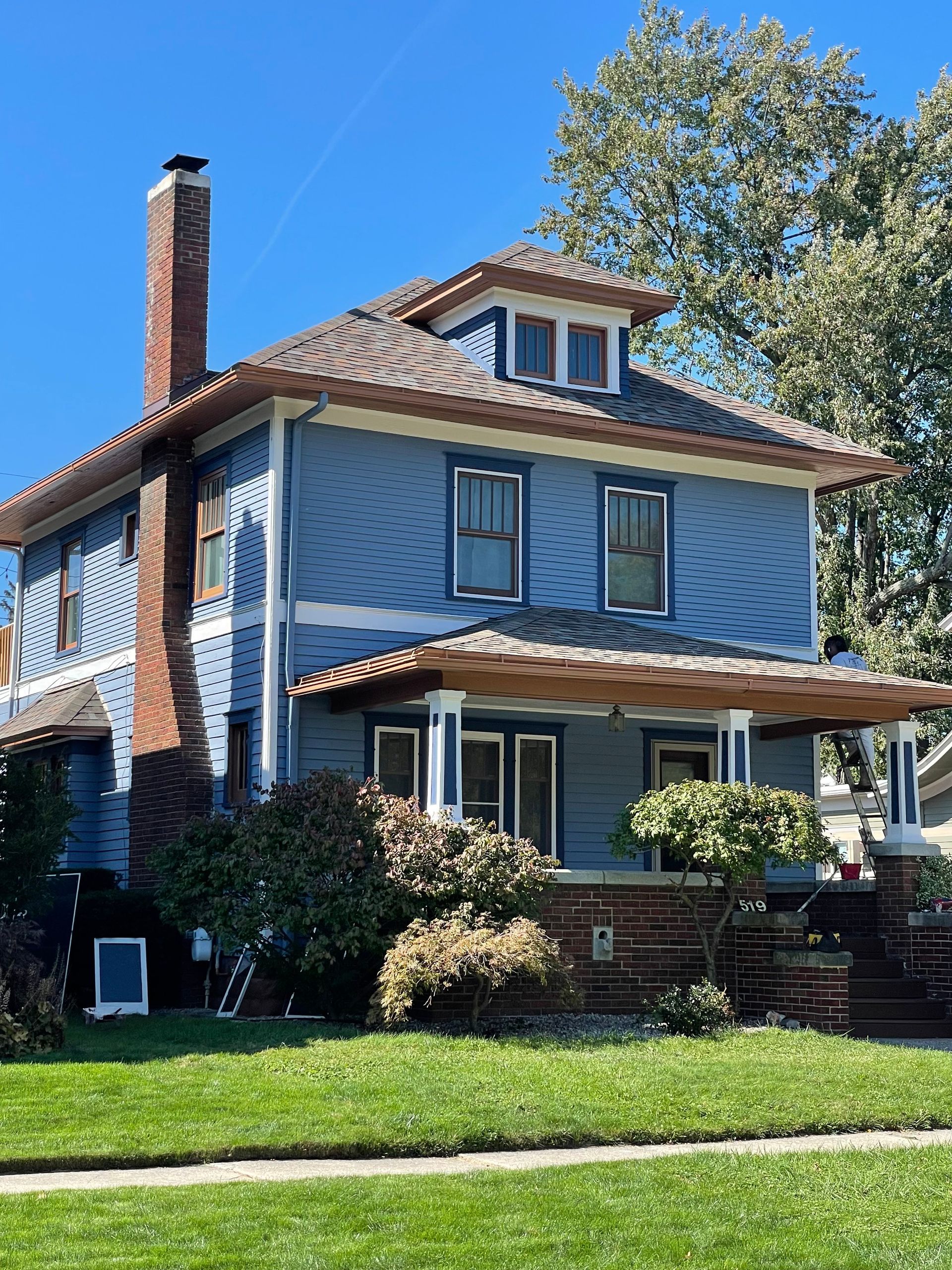 Blue two-story house with porch and chimney, set on a green lawn with a tree on a sunny day.