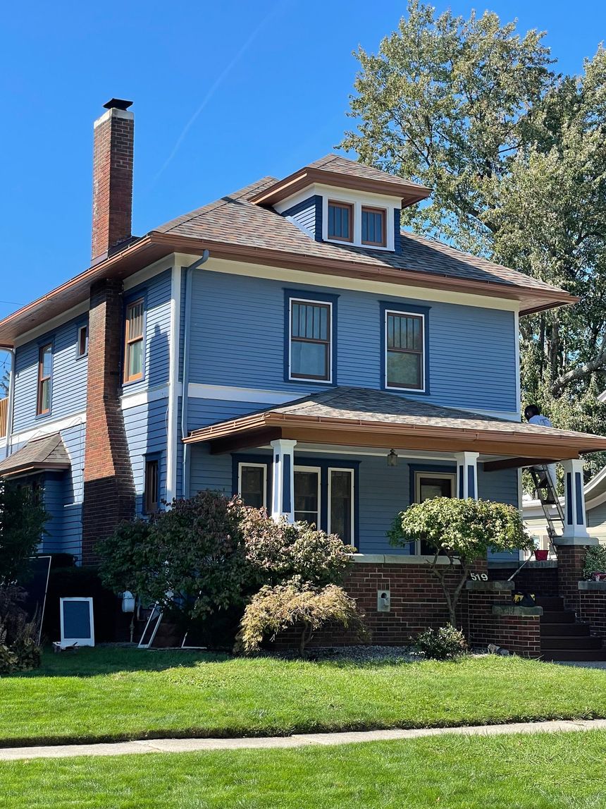 Blue two-story house with porch and chimney, set on a green lawn with a tree on a sunny day.