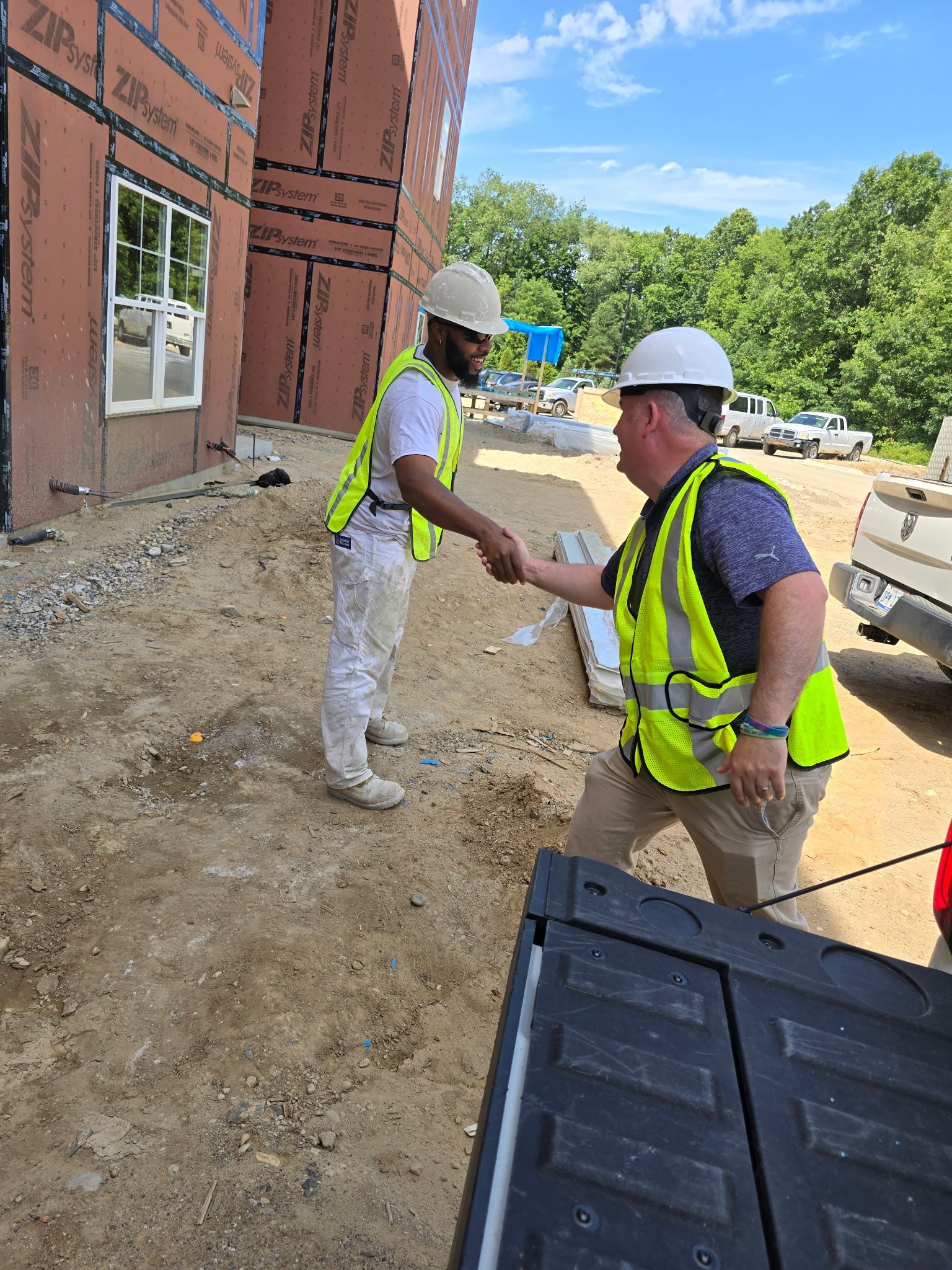 Two construction workers shaking hands at a building site; one is kneeling, both wearing hard hats and safety vests.