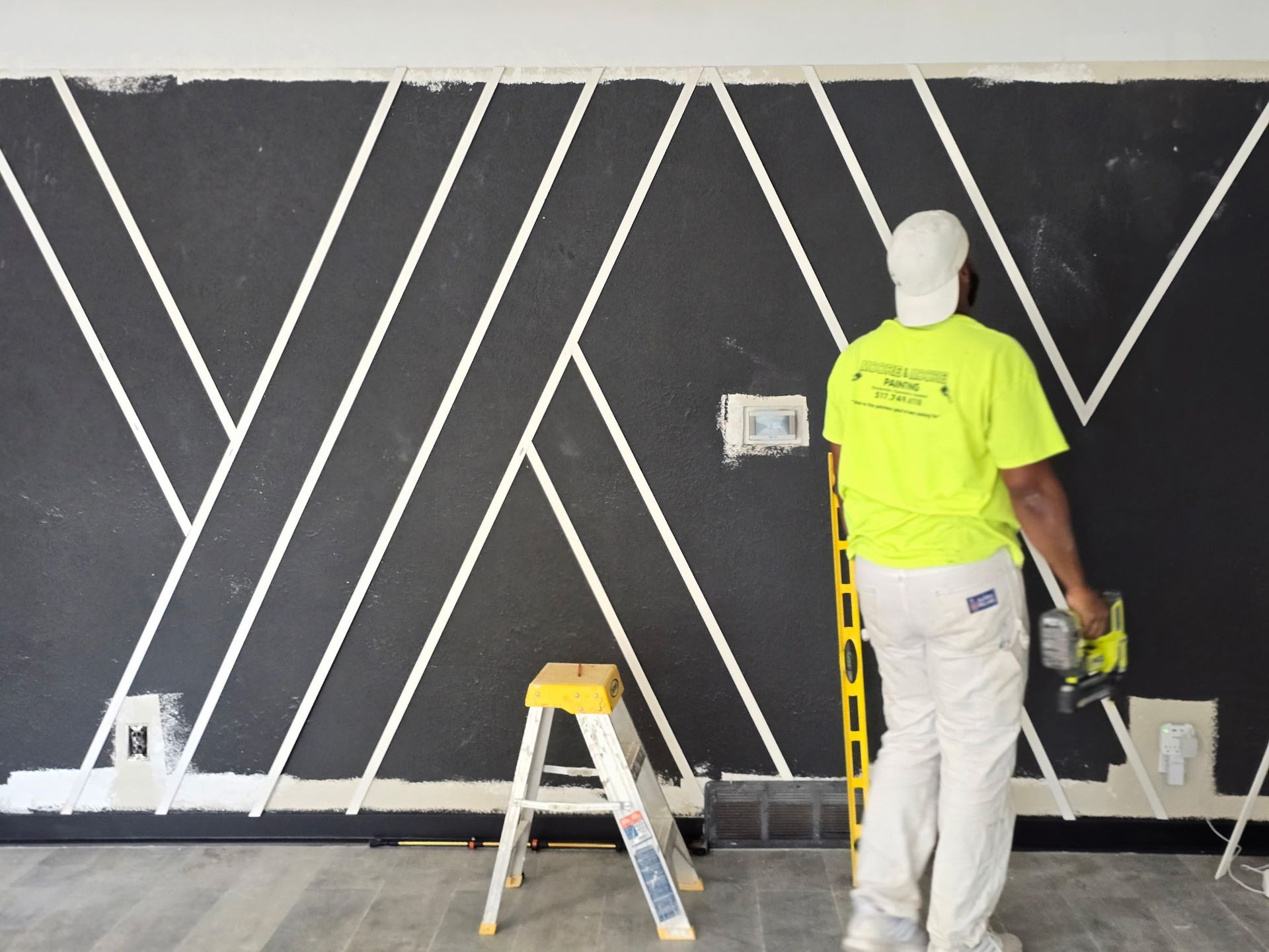 A person paints a white geometric pattern on a black wall, using a power tool, with a step ladder nearby.