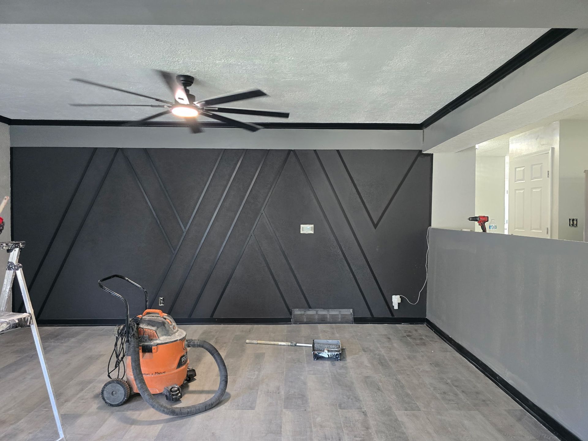 Interior with dark accent wall, grey flooring, ceiling fan, and painting tools.