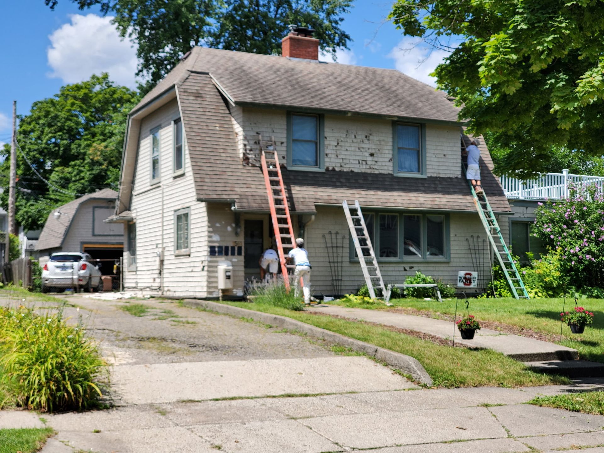 Two-story house being painted; workers on ladders. Driveway with car, trees, and blue sky.