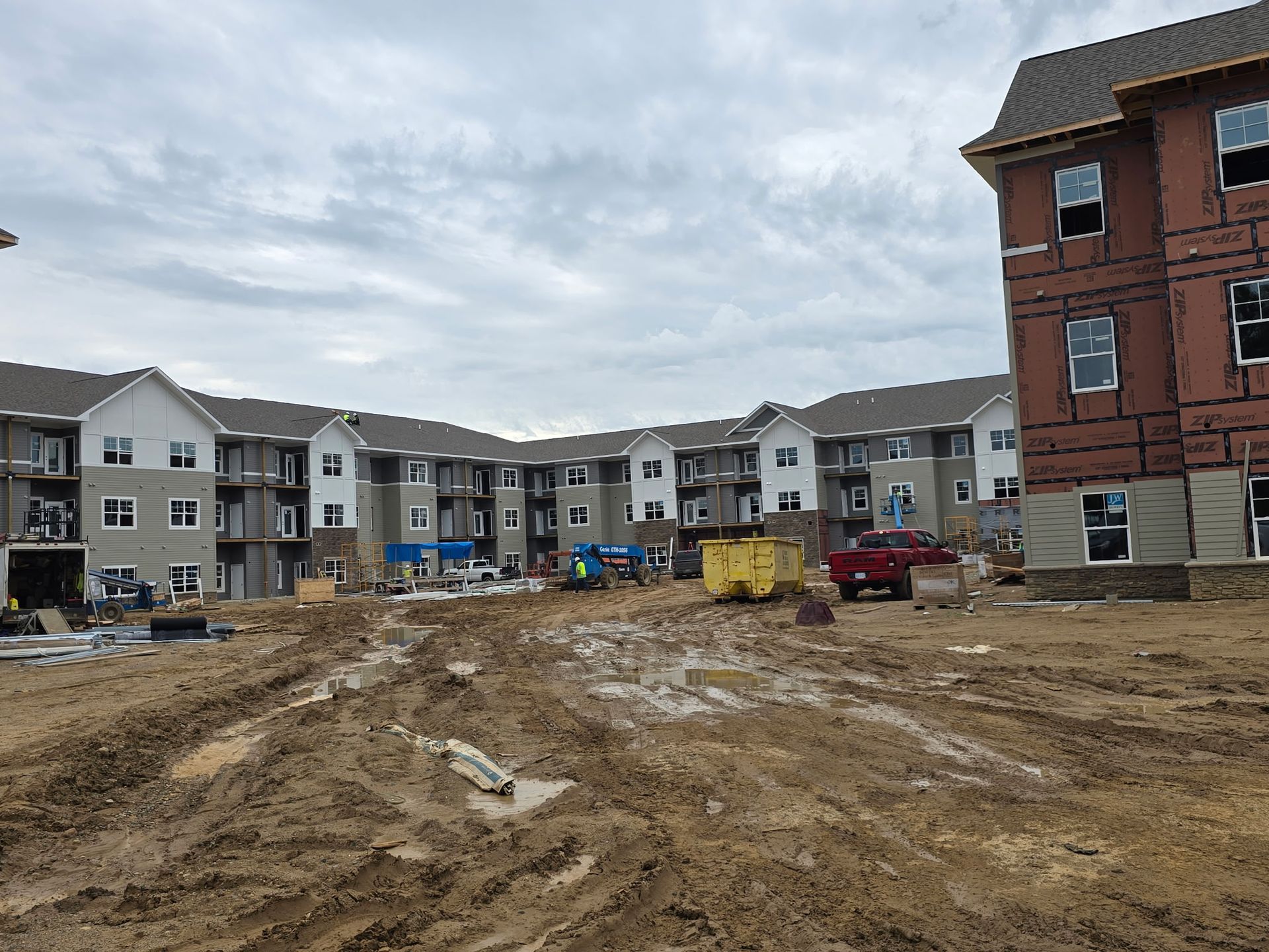 Construction site with muddy ground, multi-story buildings, and a cloudy sky.