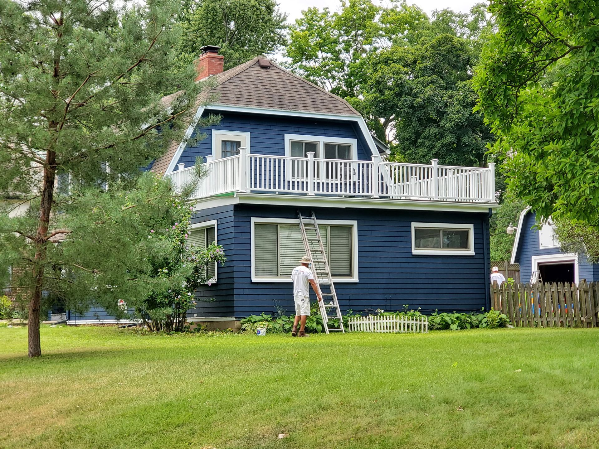 A blue house with a white balcony. A person on a ladder is working on the house. Green lawn and trees surround the house.