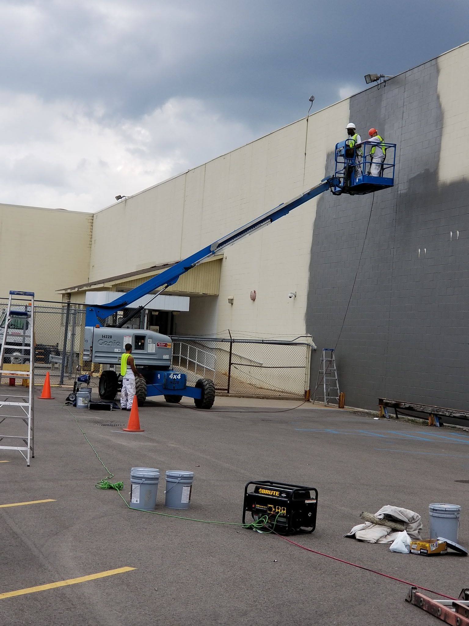 Workers in a lift painting a large building exterior gray. Blue lift, gray paint, cloudy sky.