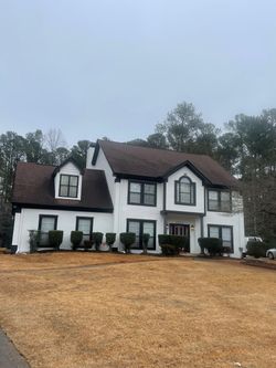 White two-story house with black trim, brown roof, and small bushes in front on a brown field.