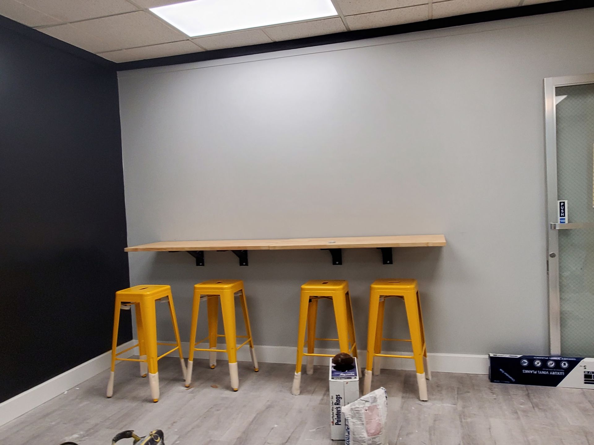 Yellow stools line a wooden countertop against a gray wall. Black wall accents and a door are visible.