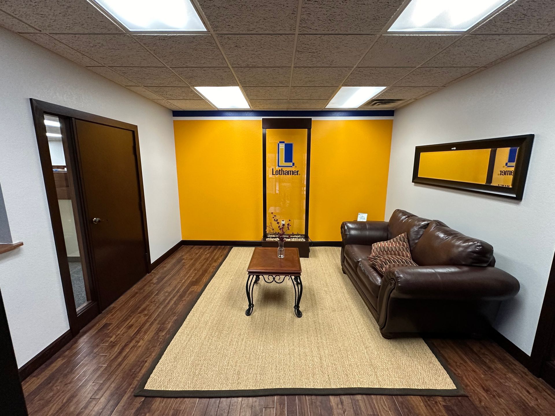 Small waiting room with brown couch, yellow accent wall, brown door, and wood flooring.