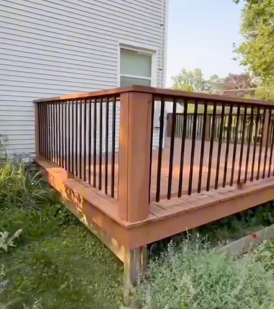Brown wooden deck with black railings next to a white house, surrounded by grass.