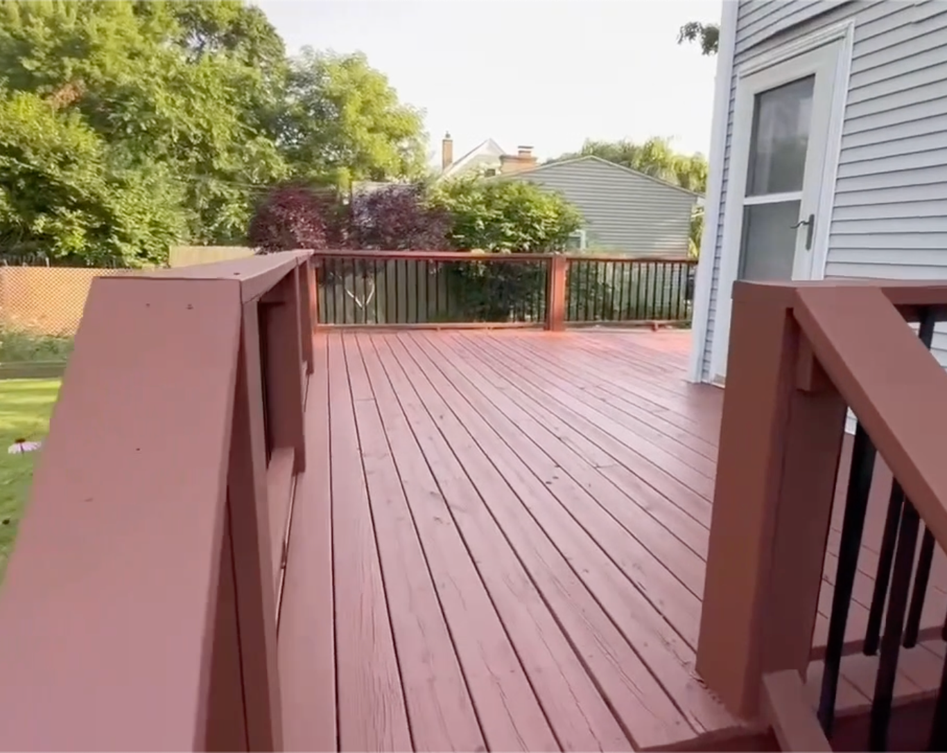 Wooden deck painted reddish-brown, with railing and a white door leading to a yard with trees.